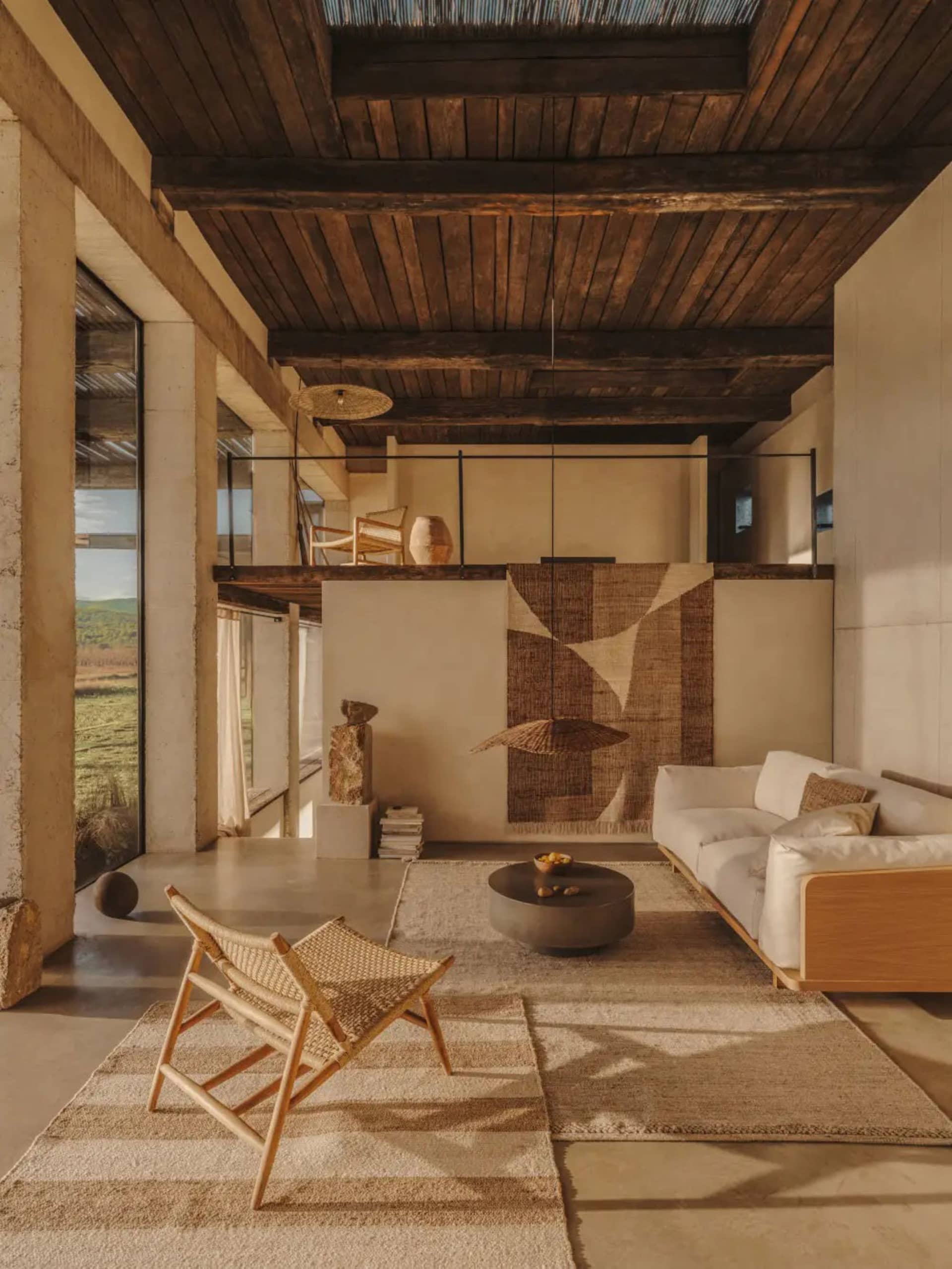 Modern living room featuring a concrete floor, wooden ceiling beams, and a textured rug, with a white sofa and woven armchair