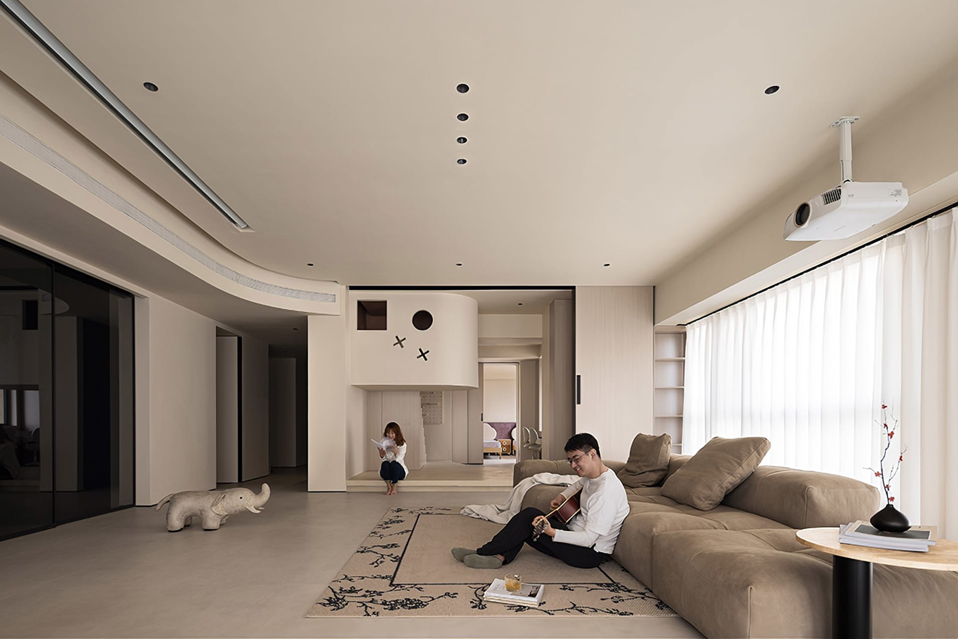 A modern minimalist living room with neutral-toned walls, furniture, and flooring, featuring a large sofa, a reading woman, a