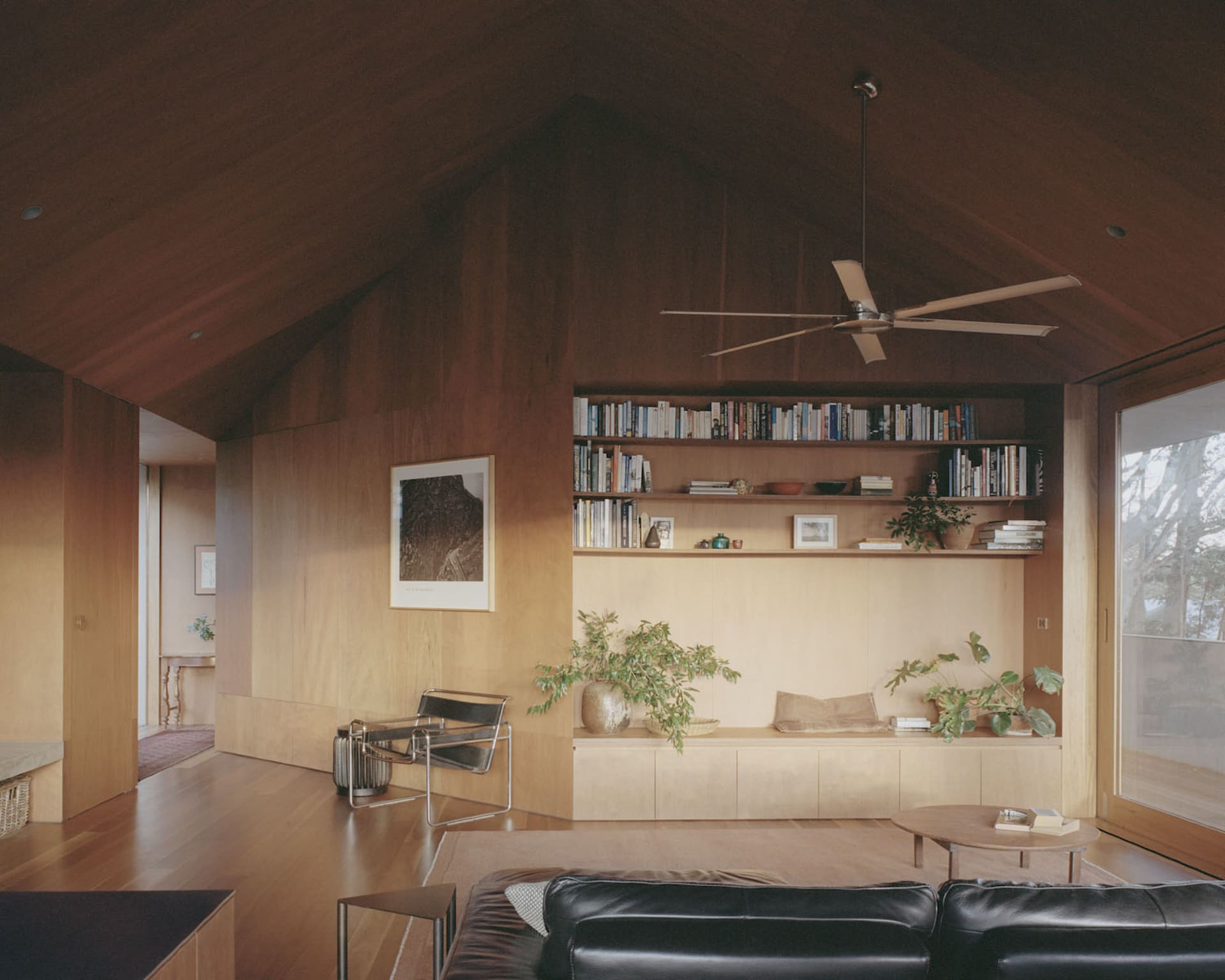 Modern minimalist living room with wooden walls, shelves, and furniture, featuring a black leather sofa, coffee table, and a