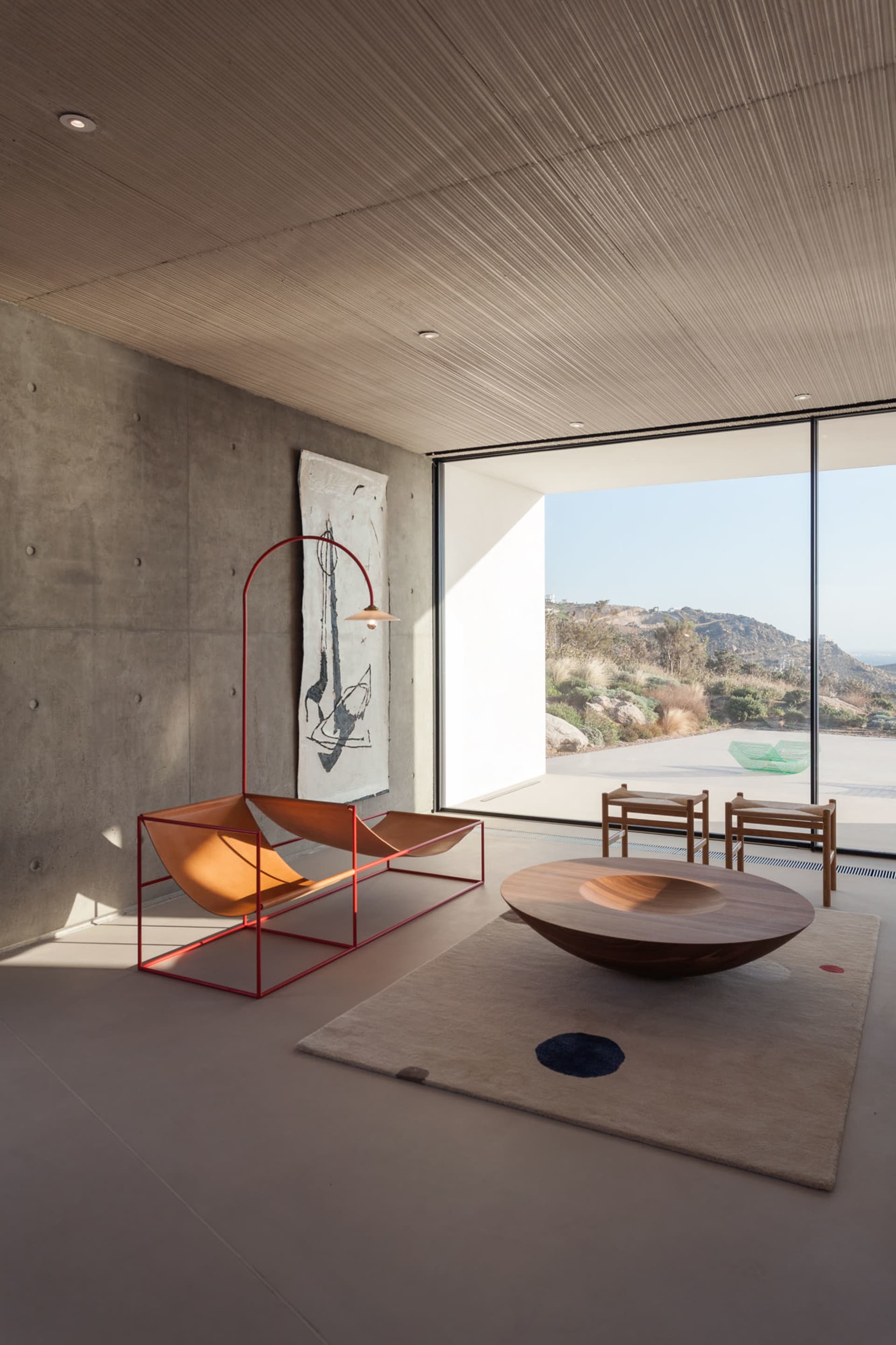 A spacious, minimalist living room featuring a concrete wall, wooden coffee table, and a large window offering a scenic view.