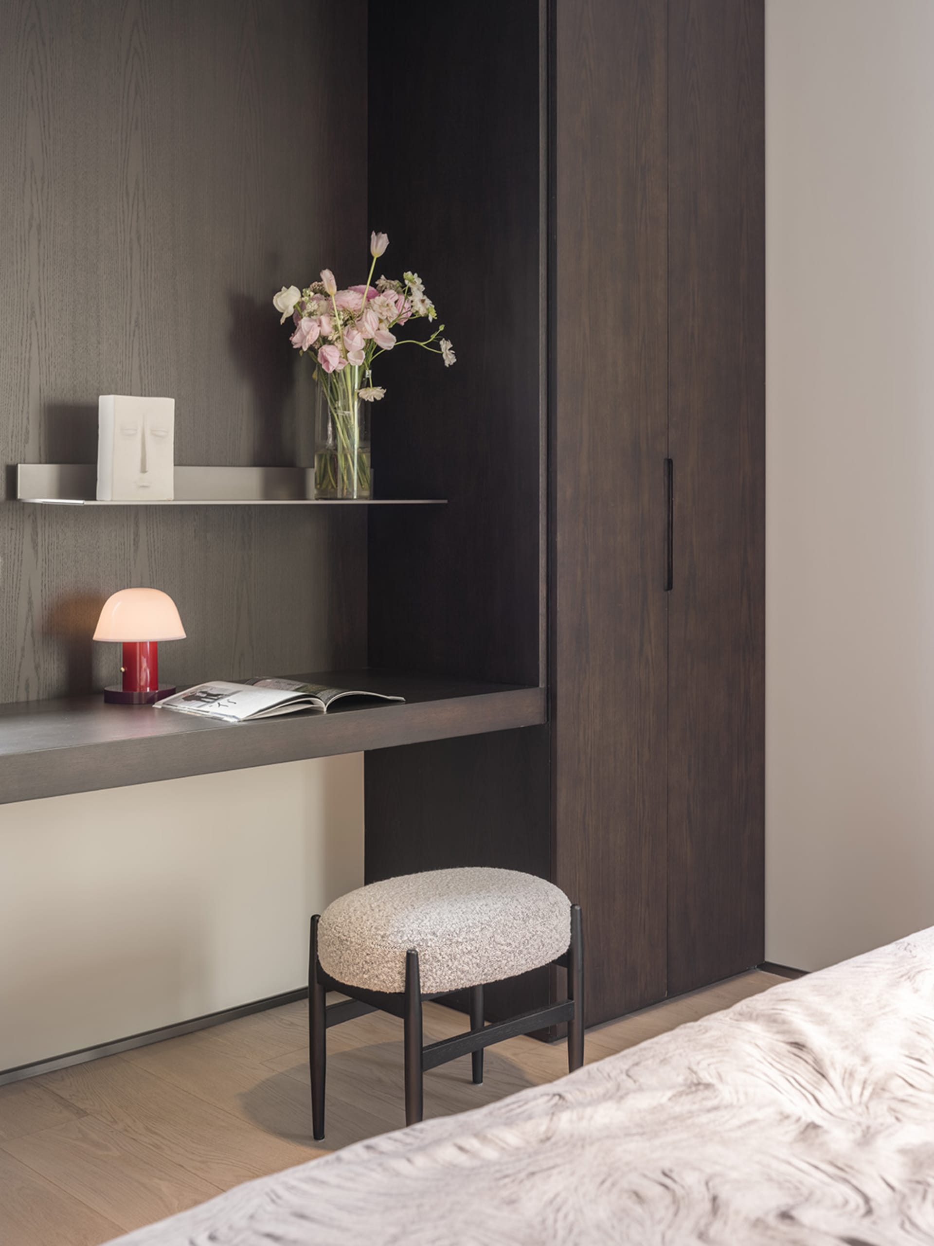 A minimalist bedroom featuring wooden furniture, a bed, a desk with a lamp and flowers, and a shelf.