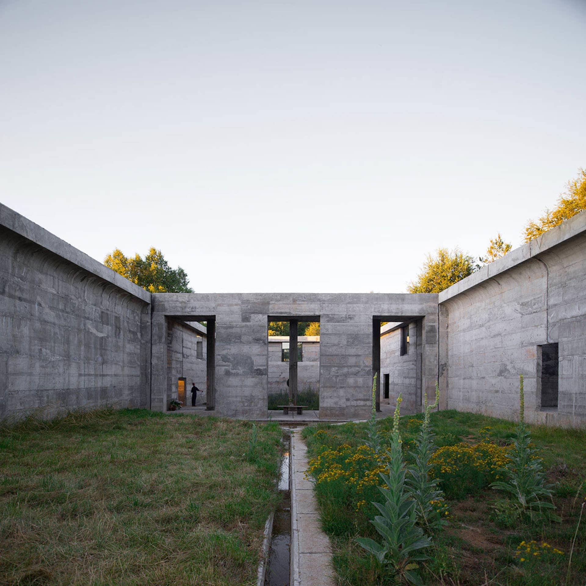 A minimalist concrete structure with large openings and a pathway leading through a garden.