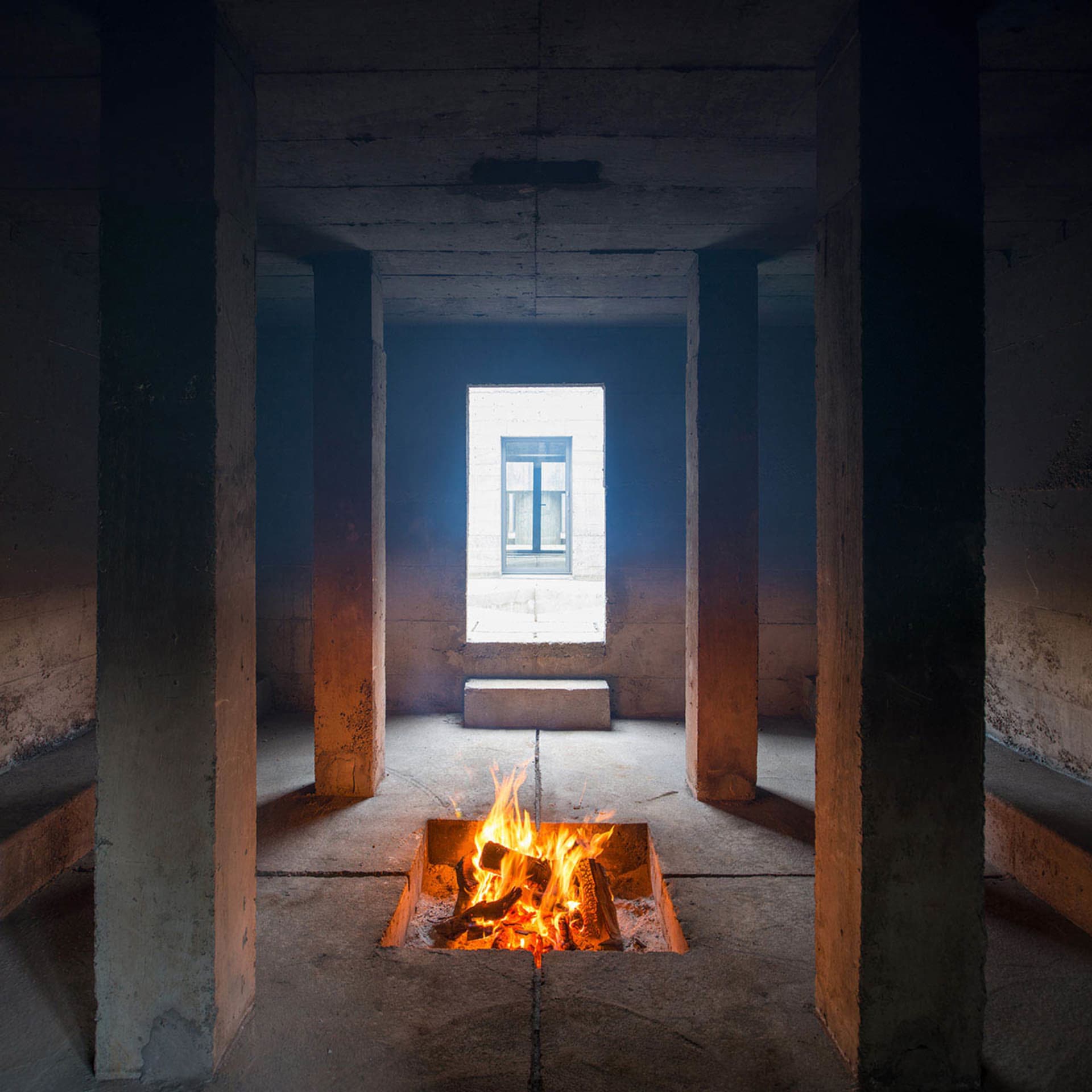 A minimalist interior with concrete walls, a burning fire pit, and a small window in the distance.