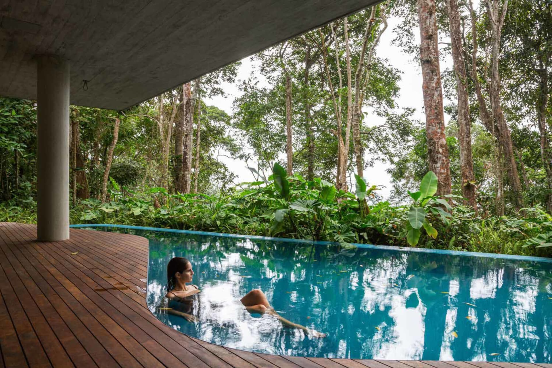 A modern poolside area featuring concrete walls, wooden deck, and blue water.