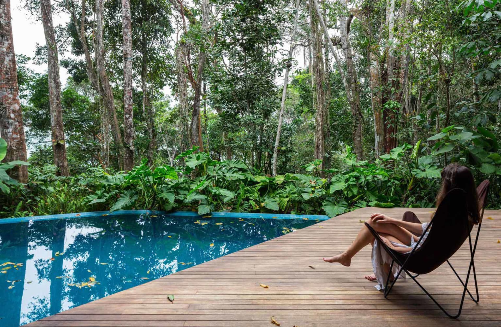 Person lounging on wooden deck overlooking lush green poolside retreat surrounded by plants.