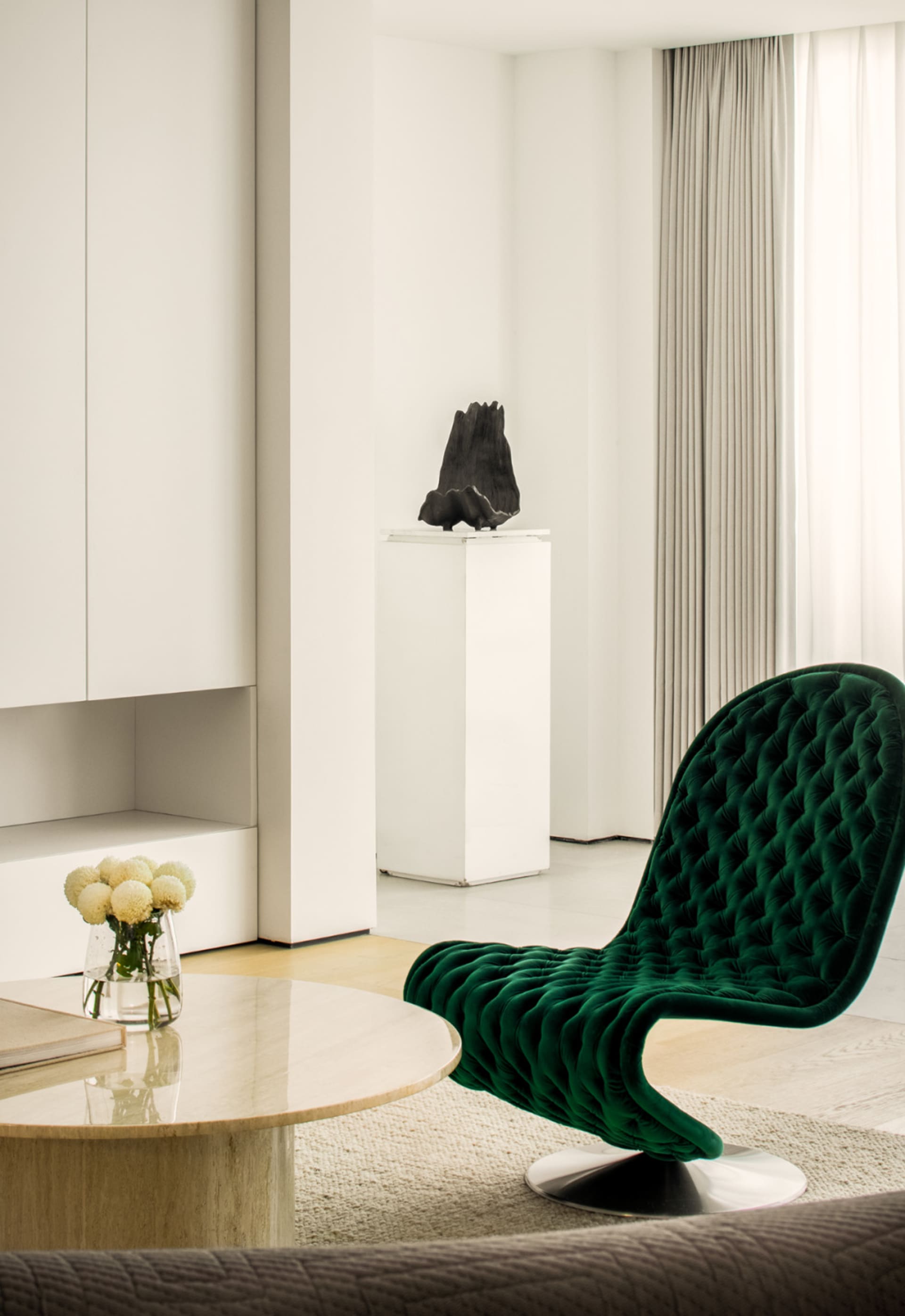 Modern living room featuring a green velvet chair with a round coffee table, beige carpet, and a sculpture on a pedestal.