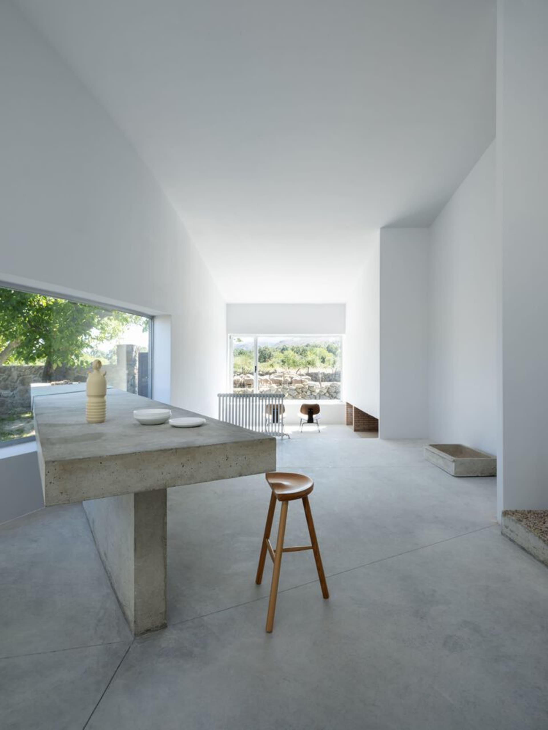 A modern minimalist kitchen featuring a concrete table and stool, surrounded by white walls and large windows with a view of