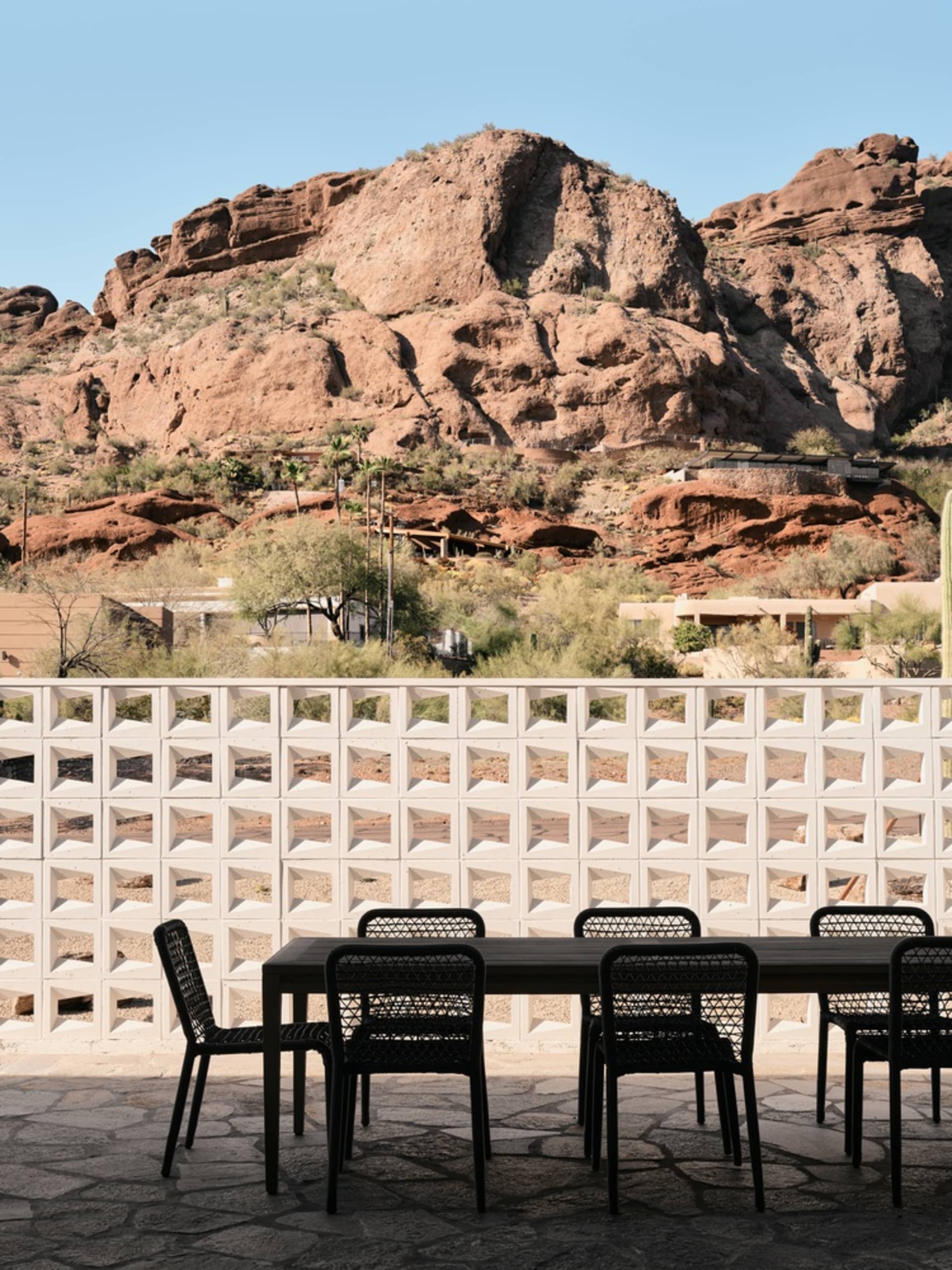 Outdoor seating area with black chairs and table in front of a white lattice wall, with rocky mountains in the background.