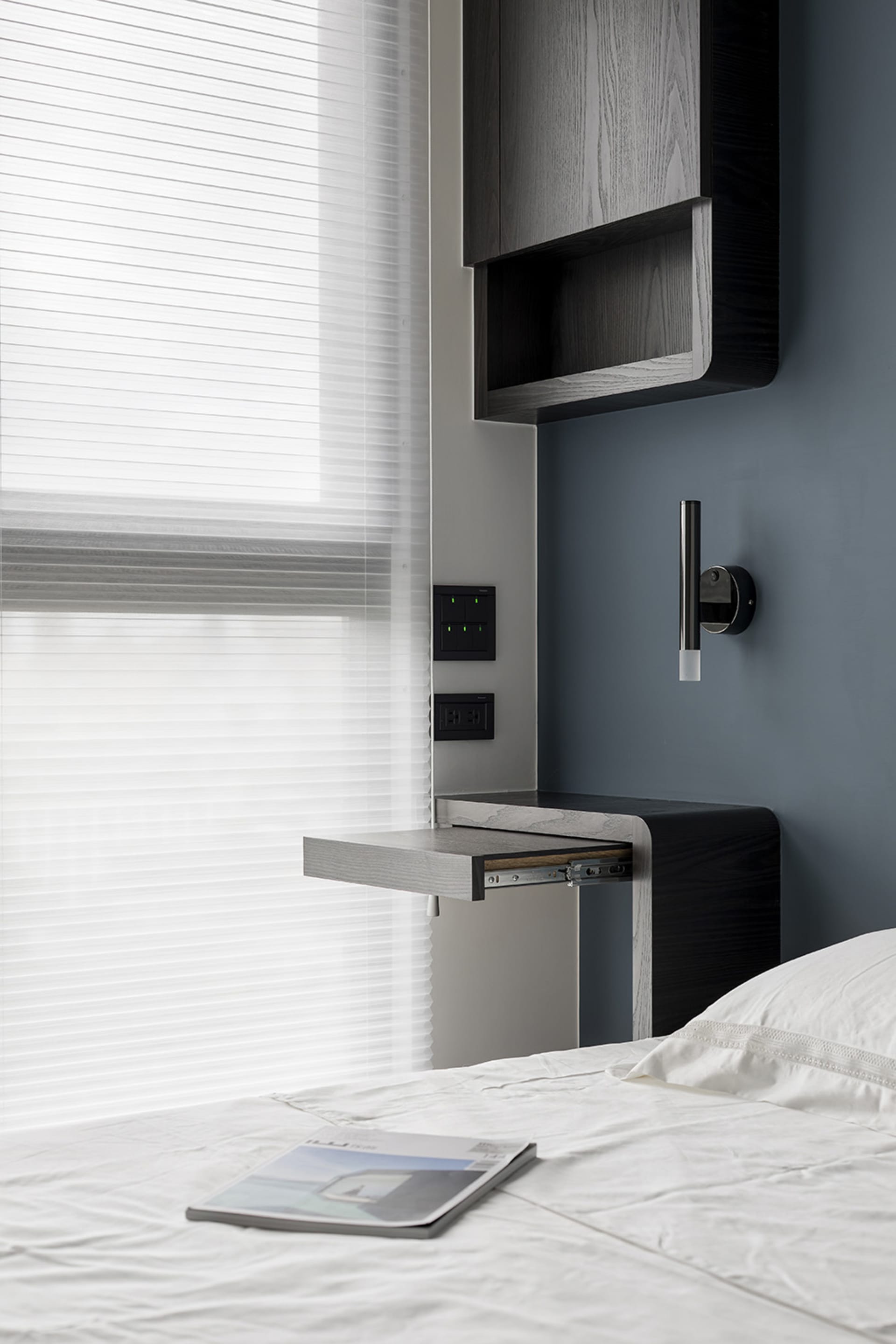 A minimalist bedroom featuring a wooden desk, blinds, and a comfortable bed with white bedding.