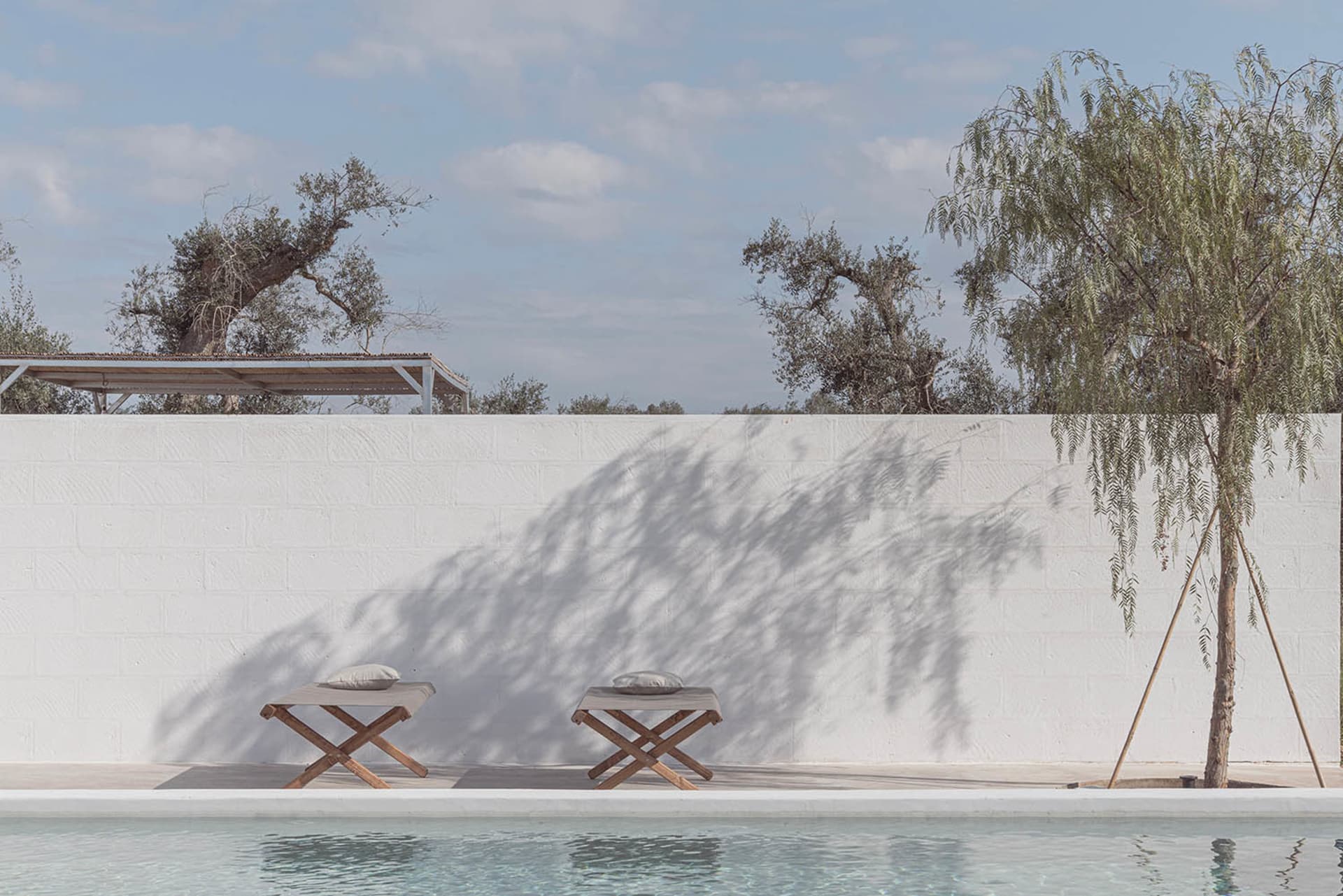 Two wooden stools with cushions on a minimalist patio with a concrete wall, shadows of trees cast on the wall, and a swimming