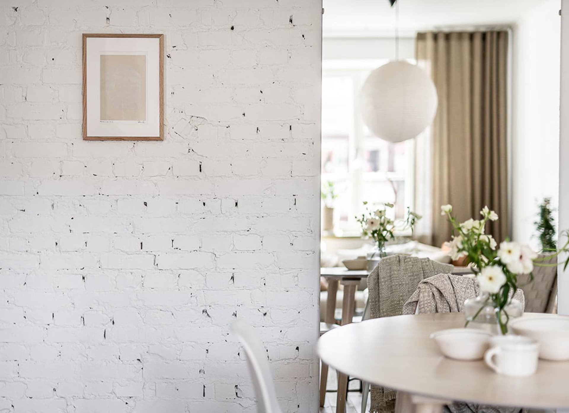 White brick wall in a minimalist dining room setting with a round wooden table, chairs, and decorative flowers.