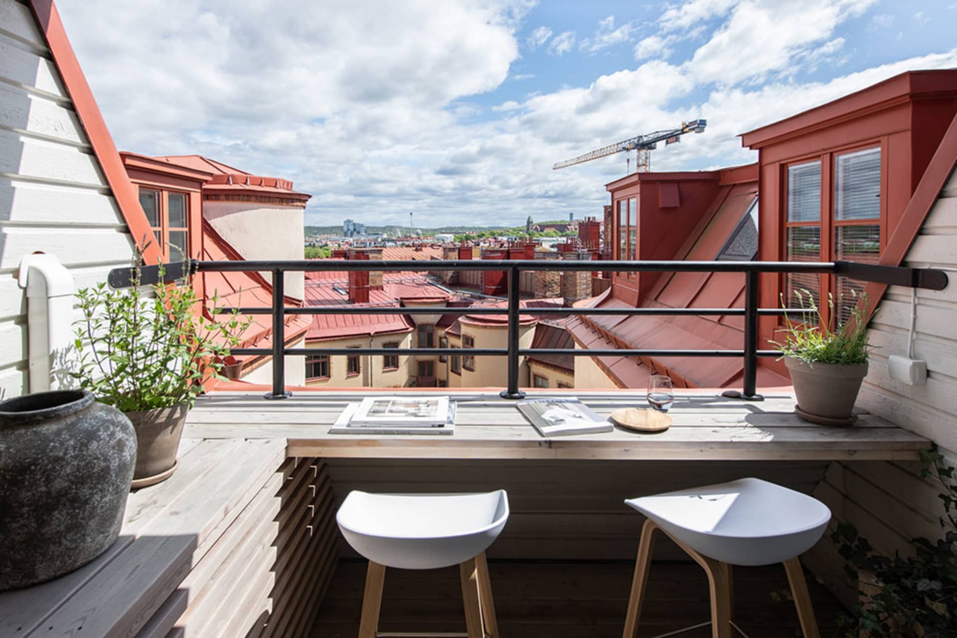 A balcony with wooden railings offering a view of rooftops and the cityscape below, with potted plants and seating.