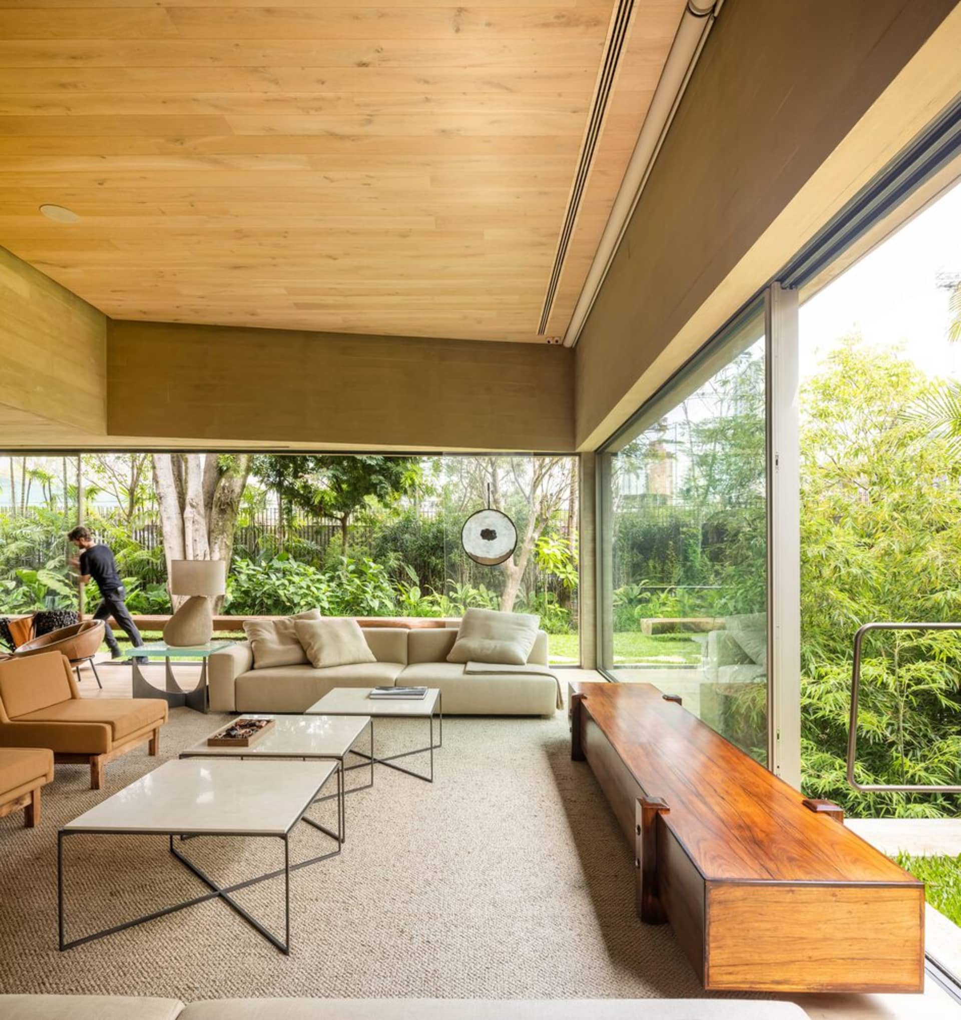A minimalist living room with wooden furniture and glass walls, featuring a view of the greenery outside.