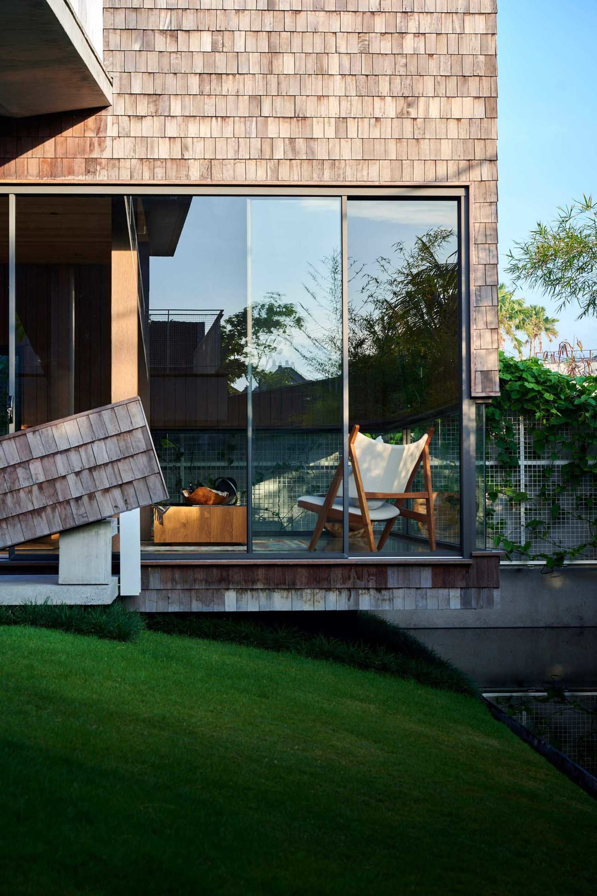 Modern residential exterior featuring a house with wood shingles, large glass window, and a white chair on the patio.