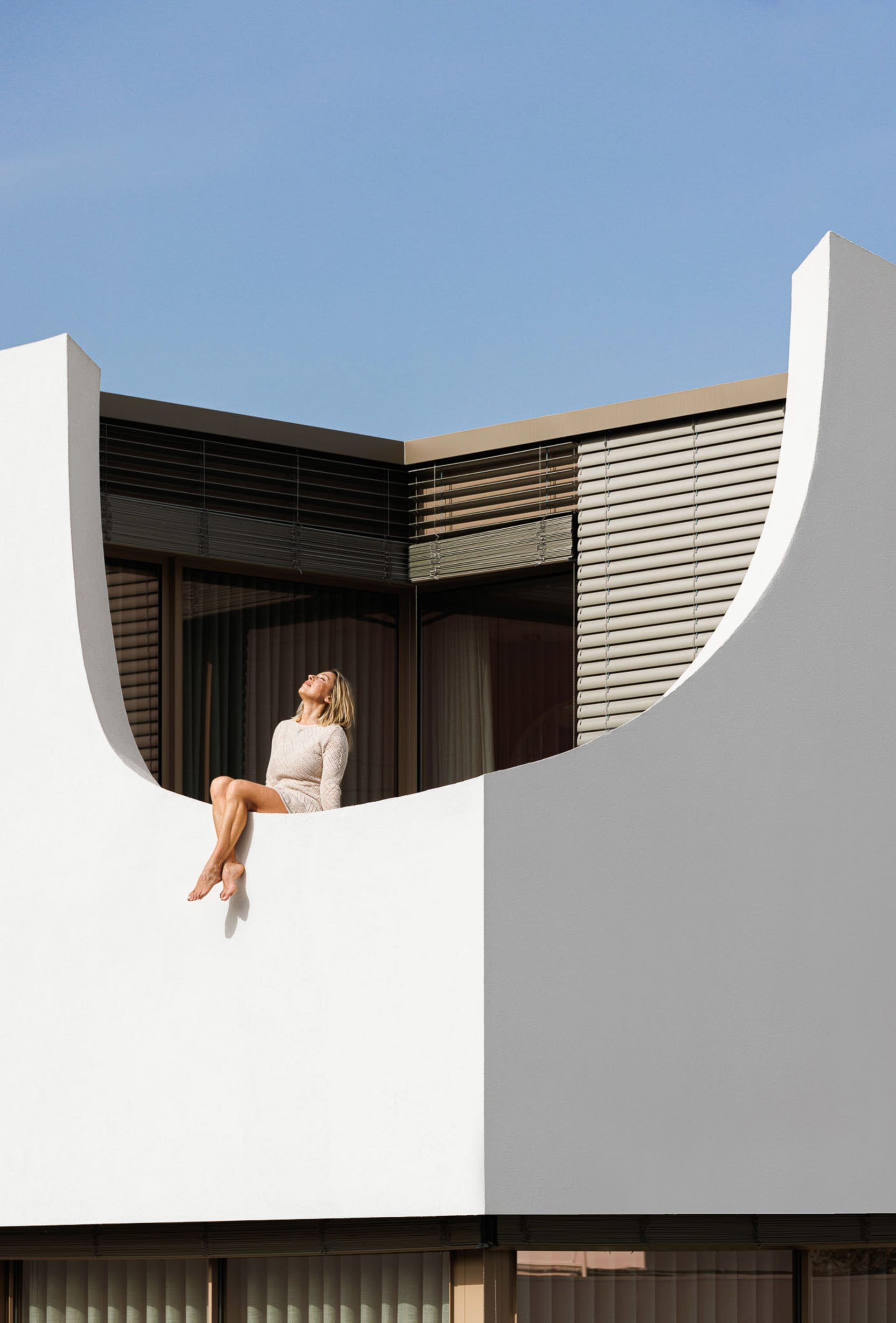 A woman sitting on a curved white architectural element against a clear blue sky.