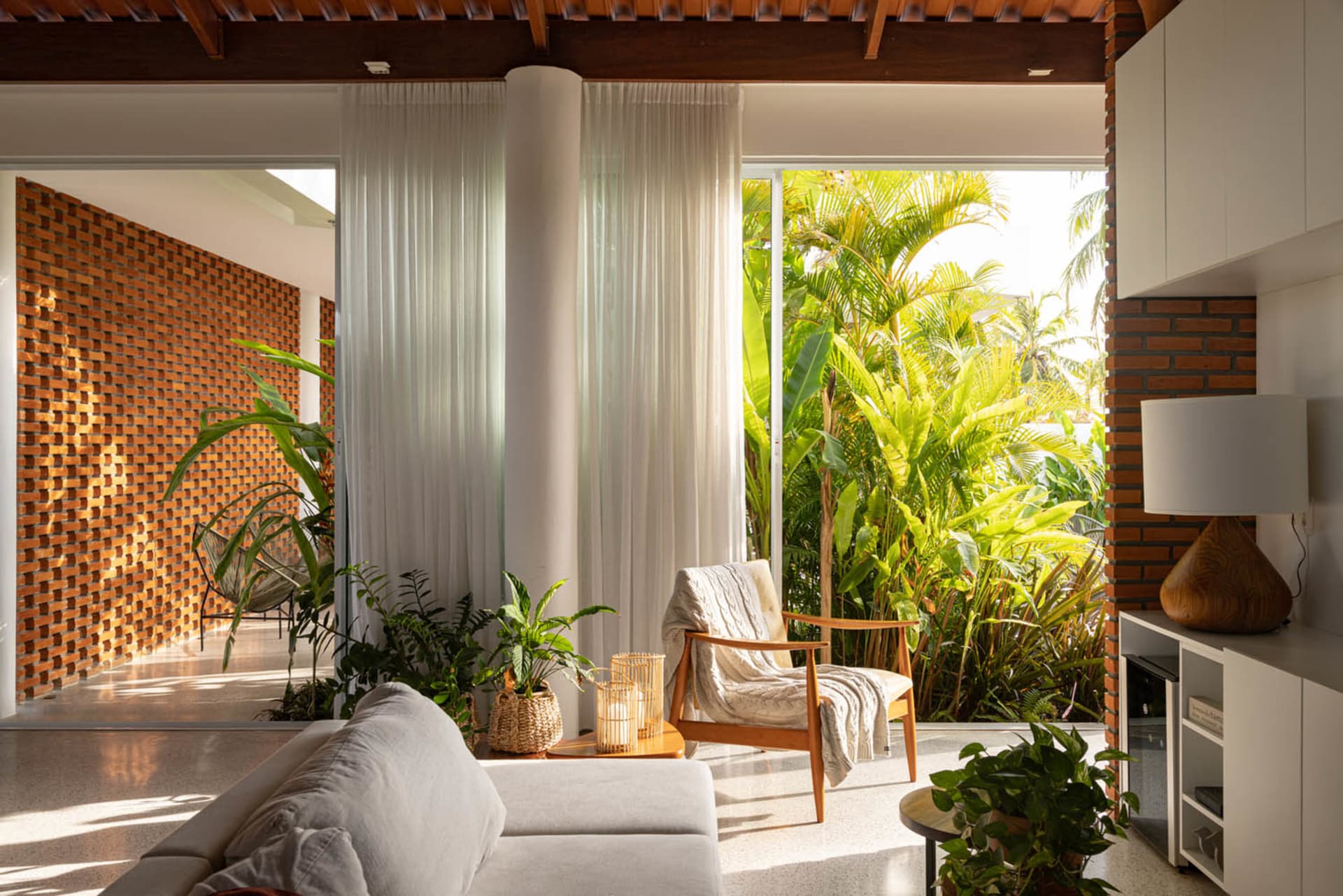 A minimalist living room with white curtains, a white fabric sofa, and wooden armchair surrounded by green plants.