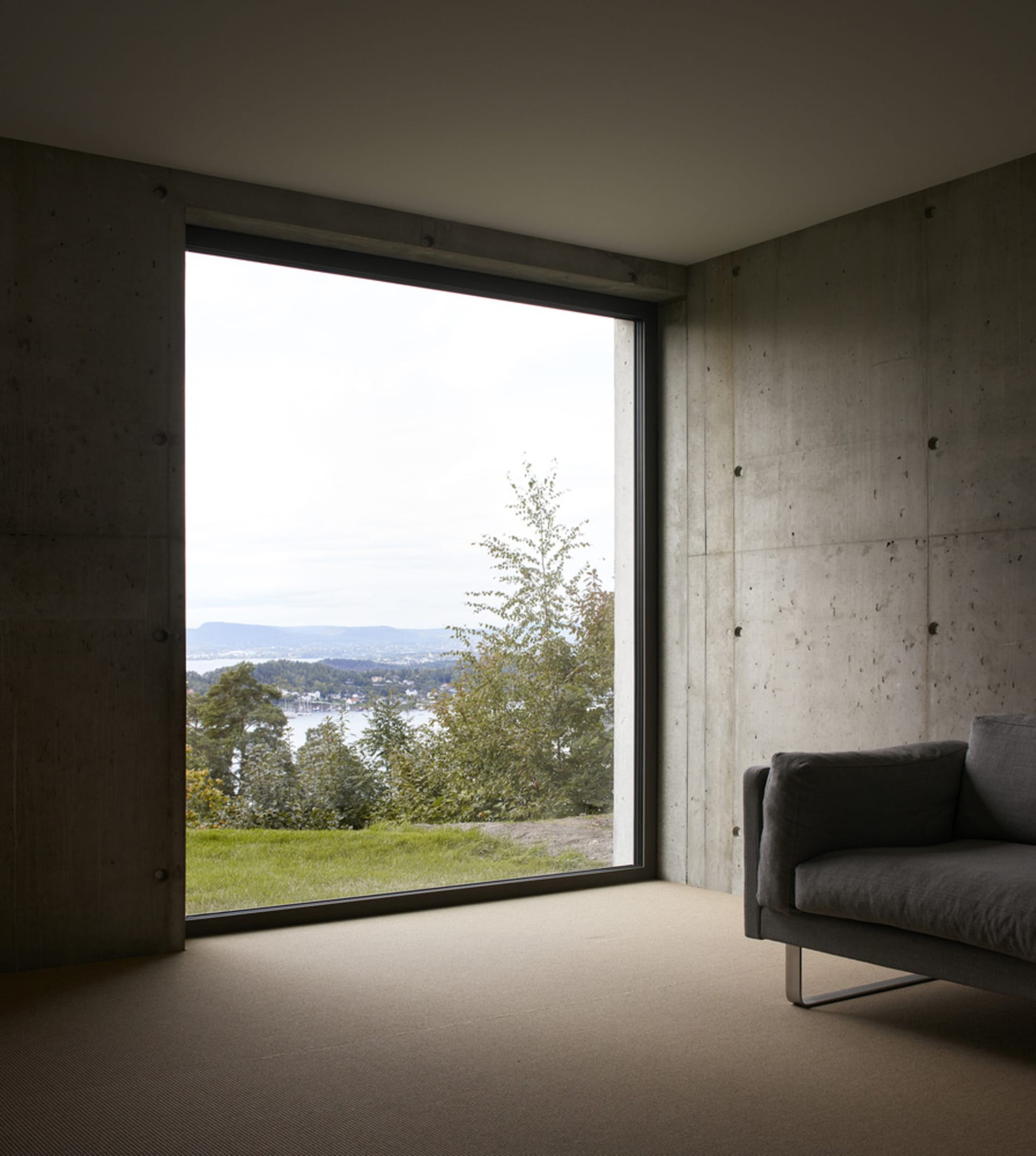 Modern, minimalist living room with a concrete wall and large window overlooking green hills and water.