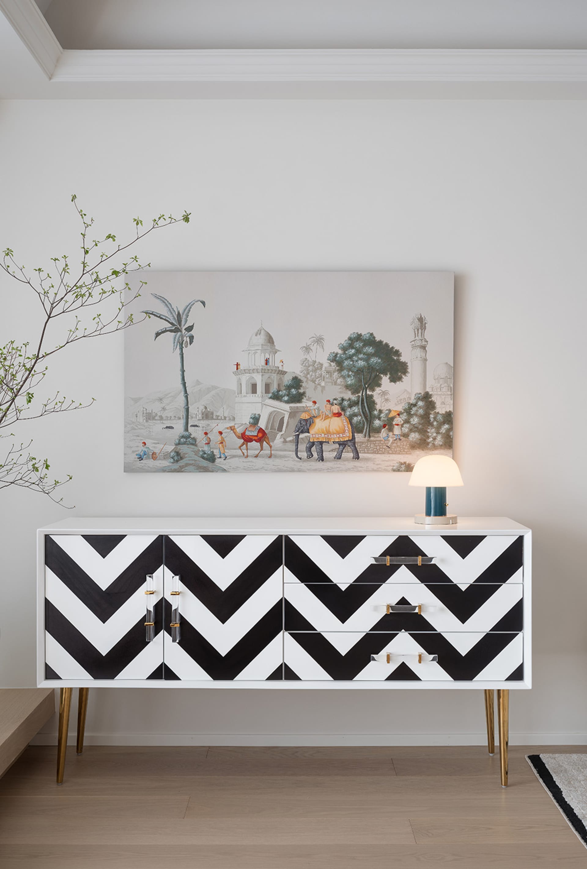A modern living room with a white and black zigzag-patterned dresser, a table lamp, and a framed painting depicting a desert
