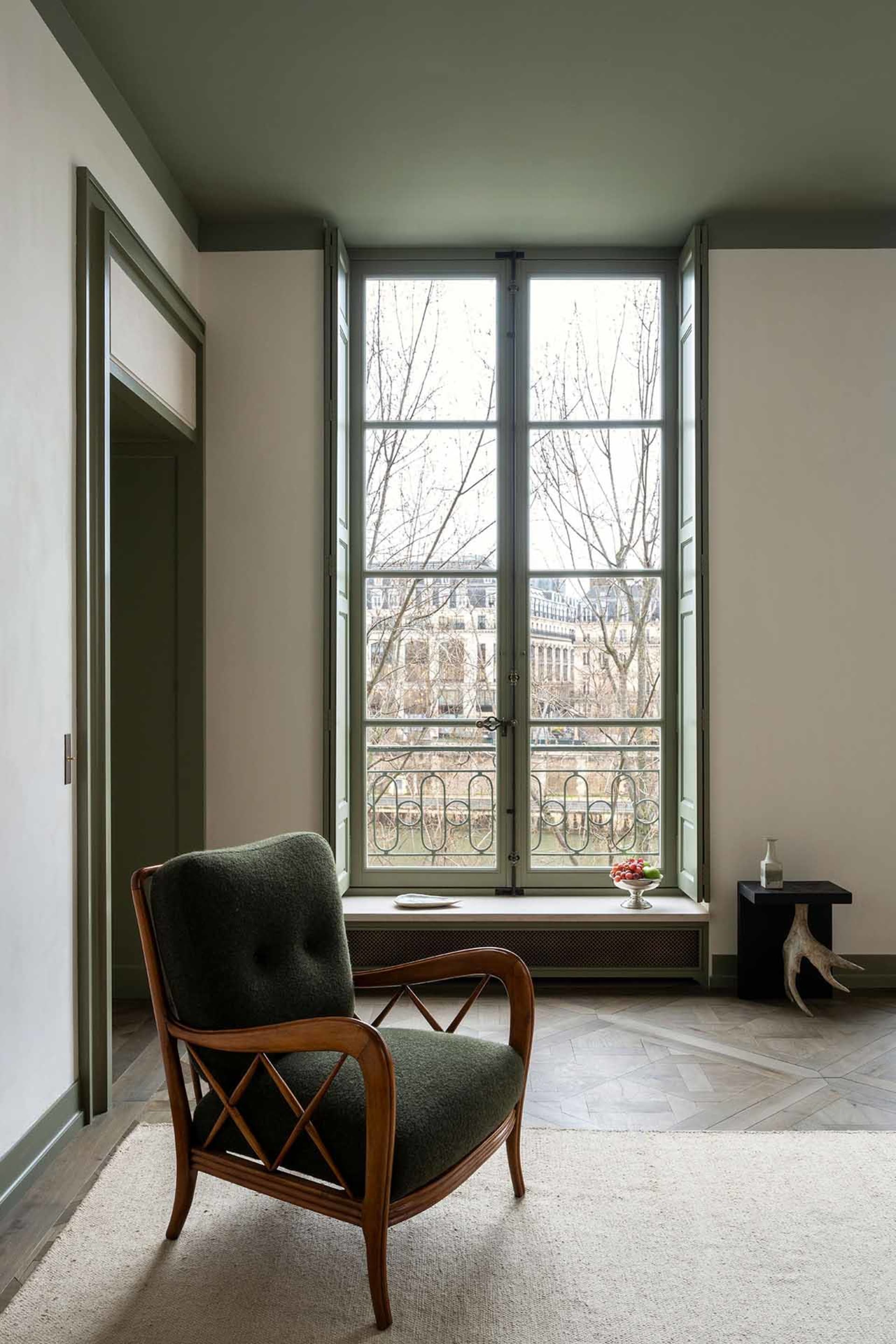 A green armchair with a wooden frame in a modern living room with large windows.