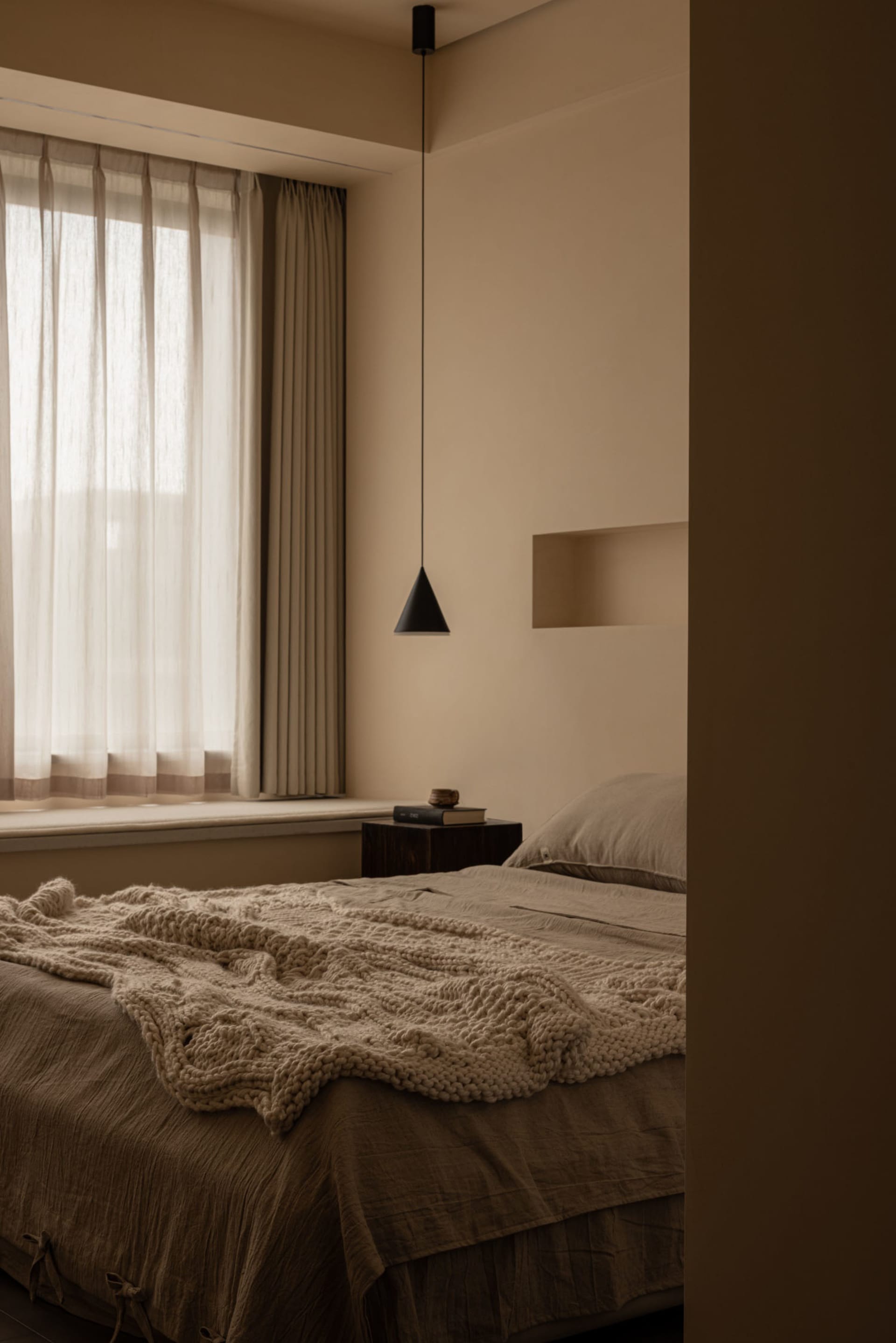 A minimalist bedroom with neutral-toned fabrics, featuring a bed, window with curtains, and a hanging lamp.