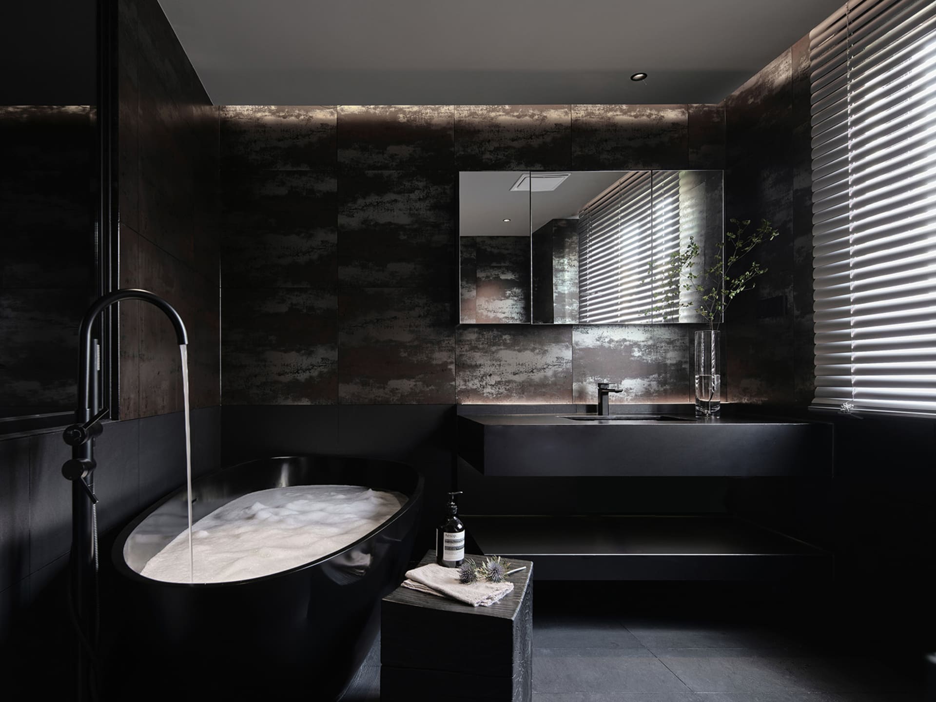 A minimalist modern bathroom featuring black tiles and concrete surfaces with a freestanding bathtub filled with bubbles unde