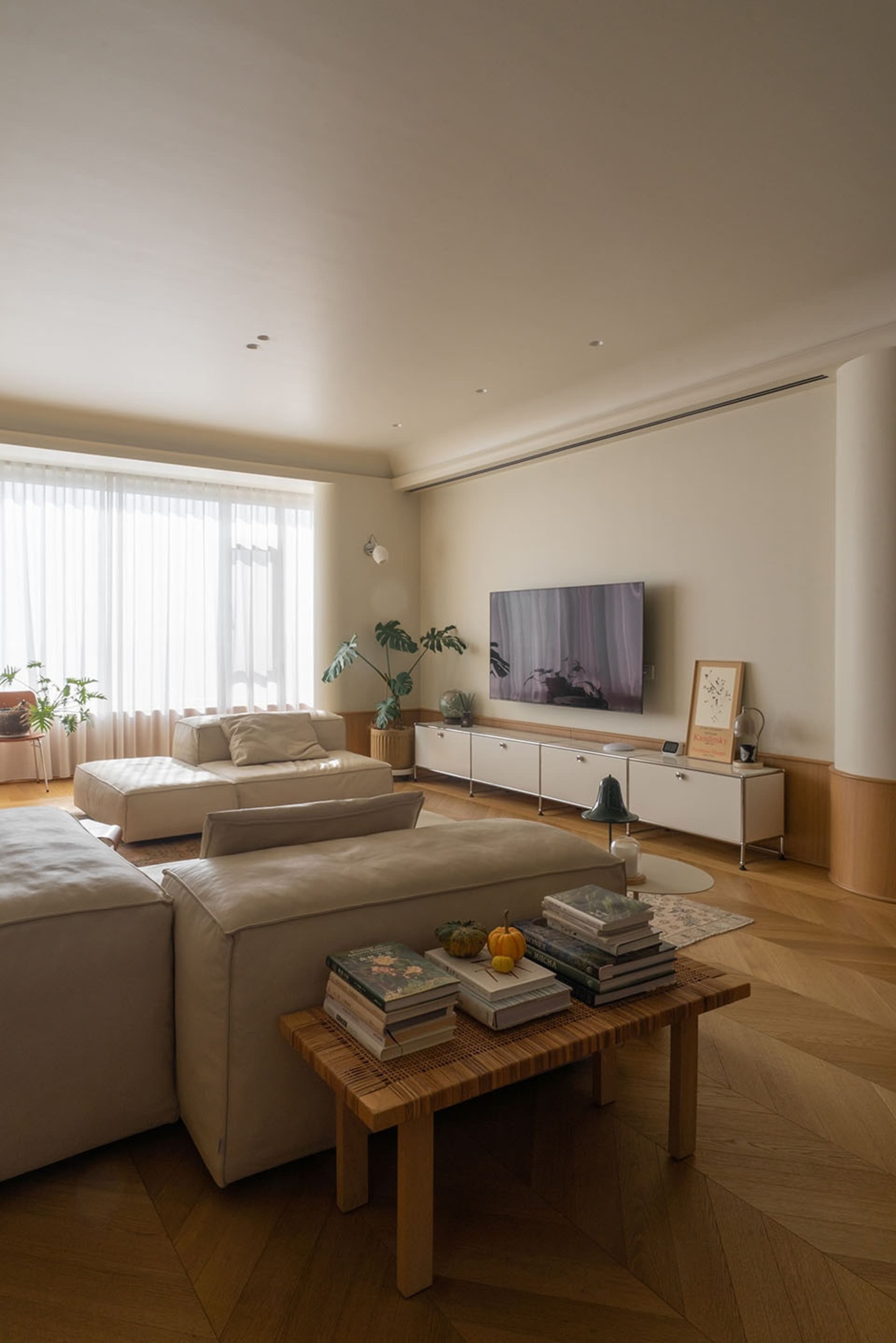 A minimalist living room featuring a beige sectional sofa, a wooden coffee table with books and decorative items, and a white