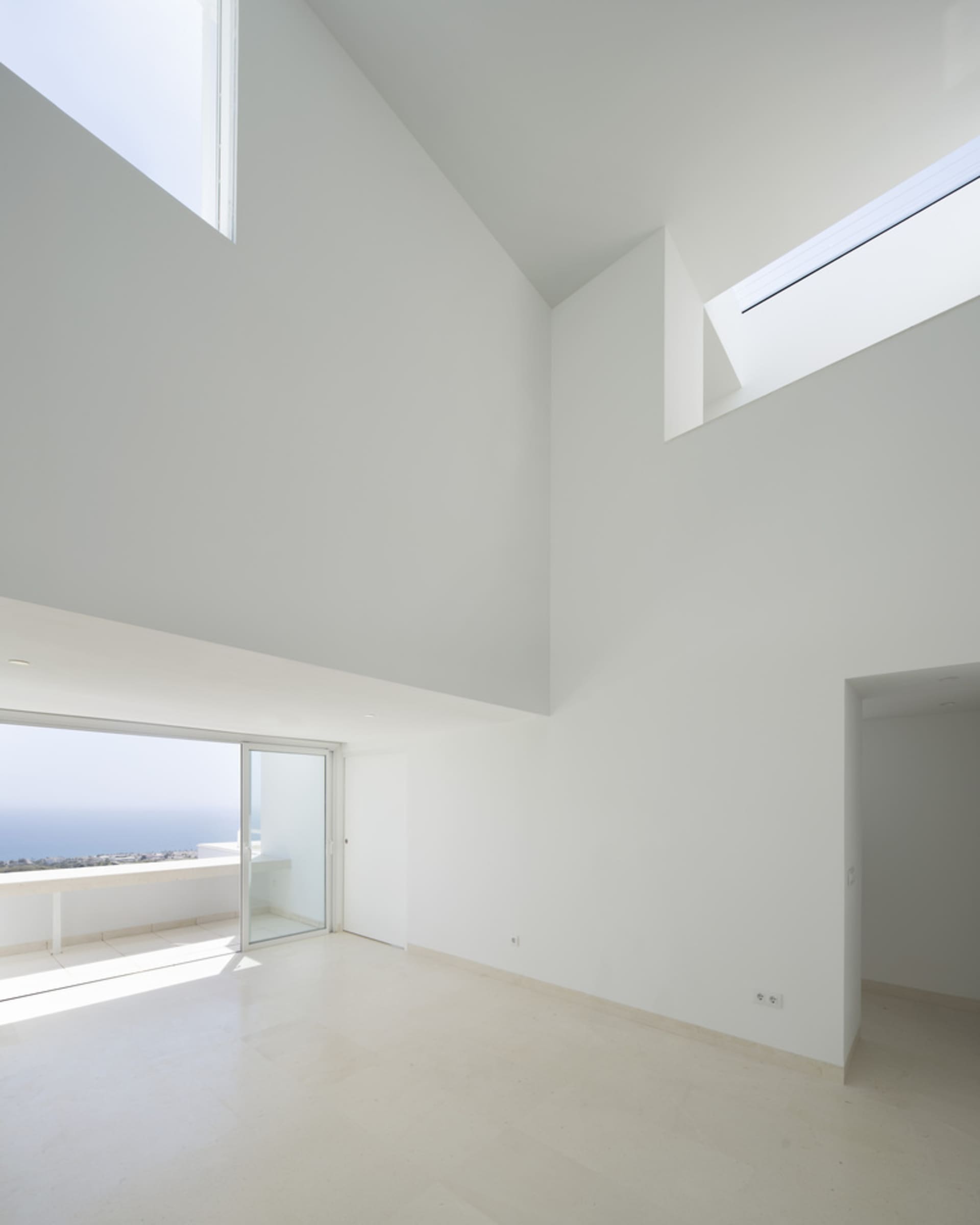 An empty minimalist living room with high ceilings, large glass windows overlooking the ocean, and white walls and floor.