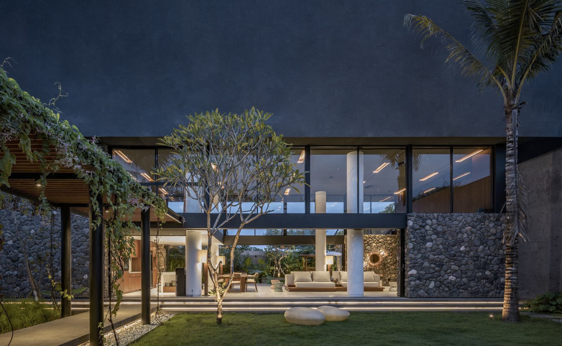 A modern house with concrete walls, glass windows, and stone accents illuminated during night time.