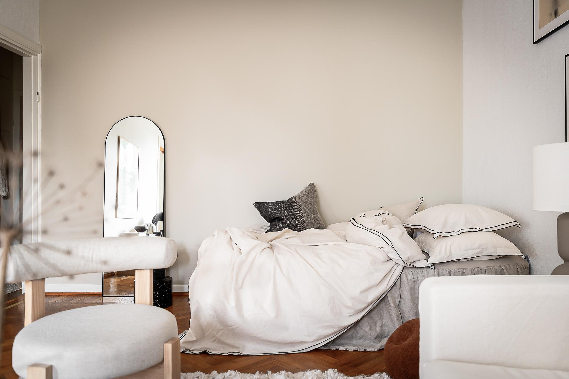 A minimalistic bedroom with a bed covered in white linen, wooden furniture, and a large mirror on the wall.