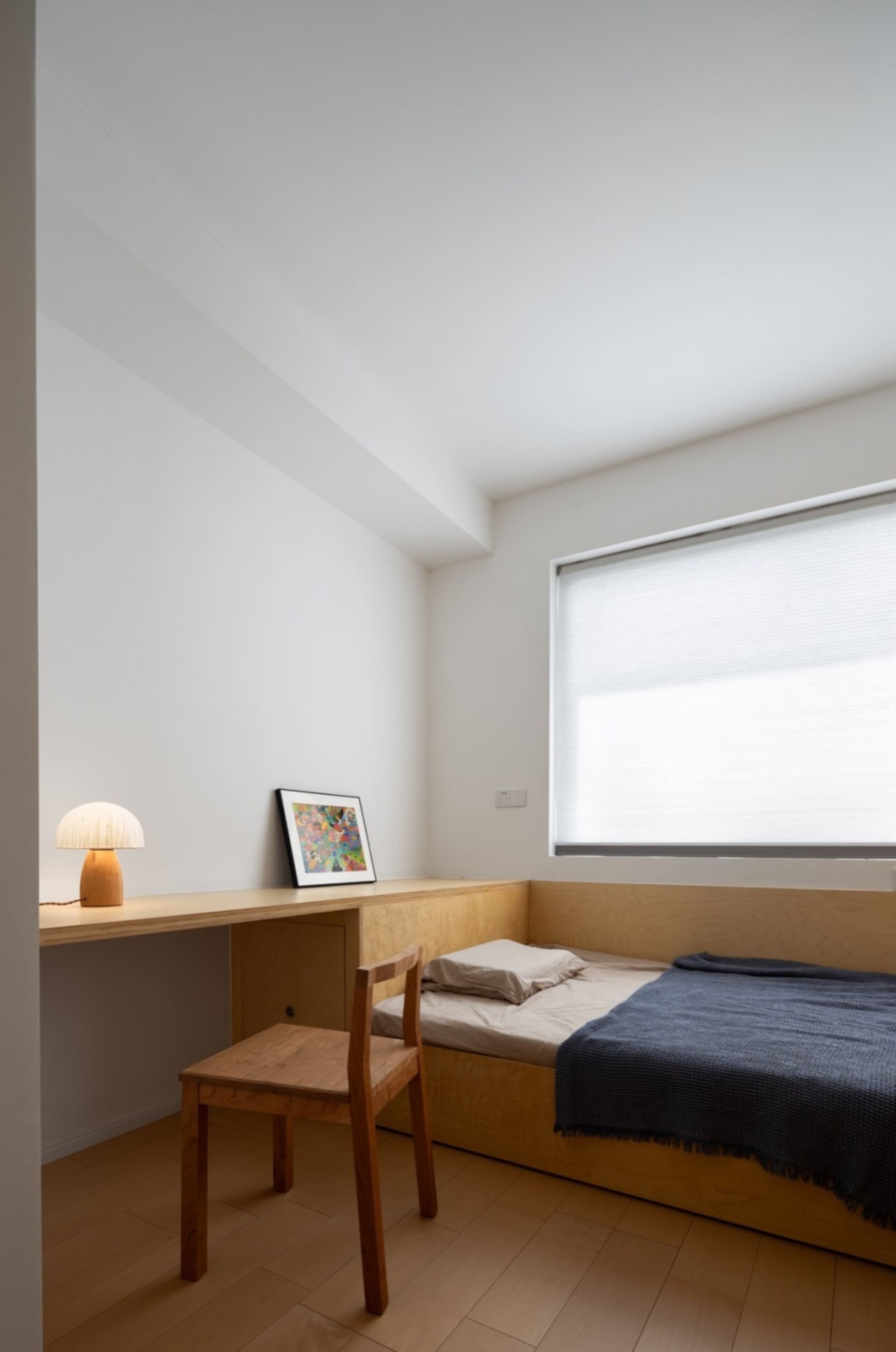 Modern minimalist bedroom with a solid wood bed, desk, and chair, featuring a large window with blinds.
