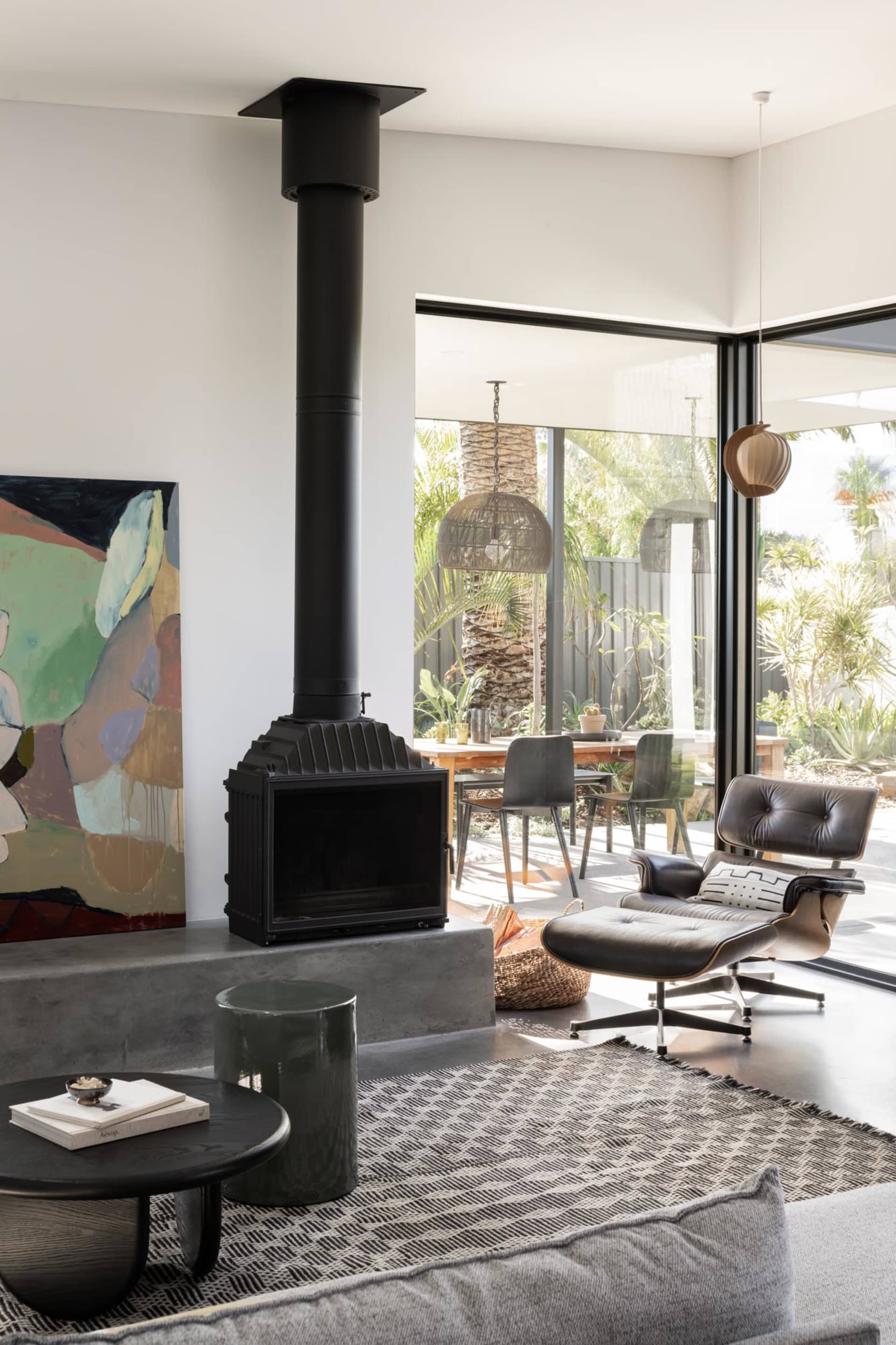 Modern industrial living room featuring a fireplace, leather chairs, concrete floor, and large windows with a garden view.