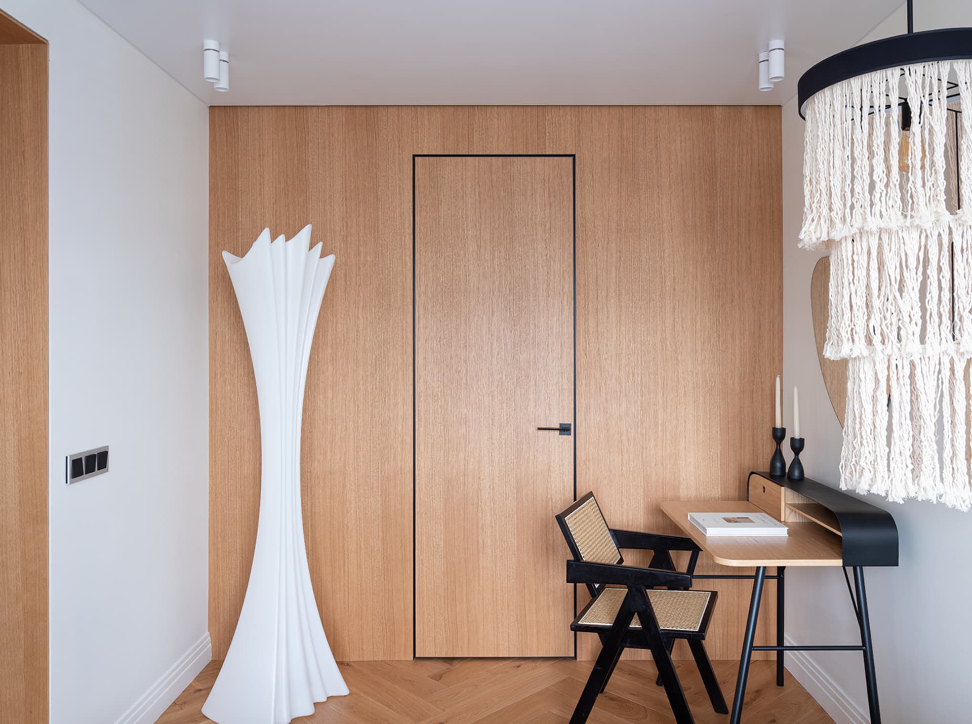 A modern home office features a wooden wall with a closed door, a sleek desk, and a minimalist white floor lamp.