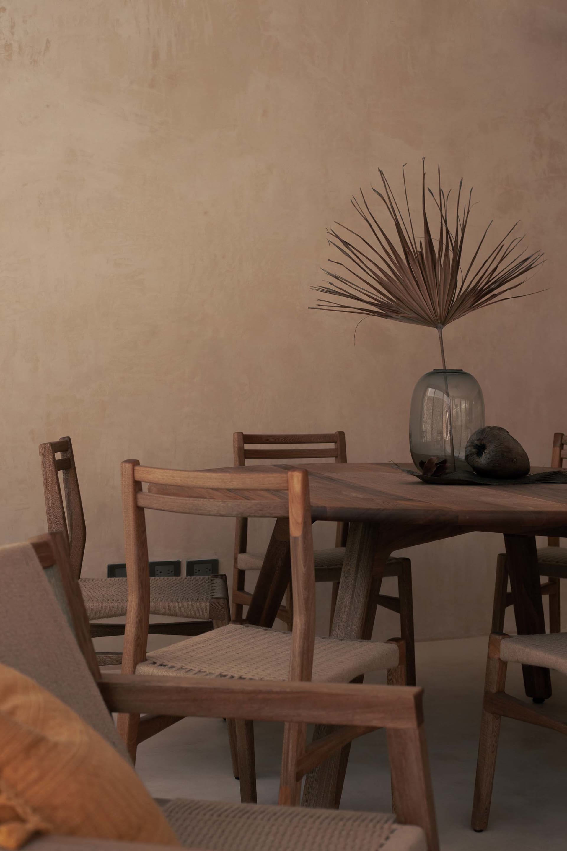 Industrial-style dining room with a round wooden table, woven chairs, and a vase with dried palm fronds.