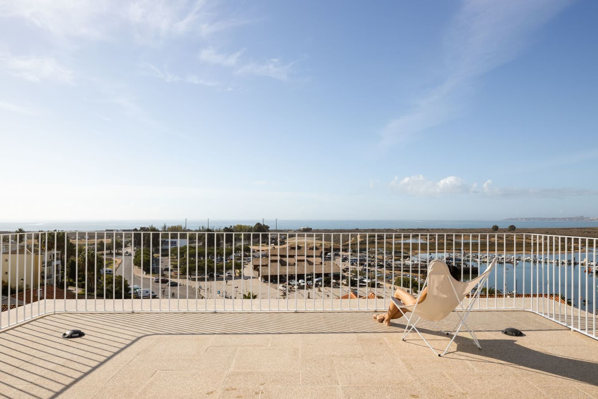 A person sitting in a white chair on a balcony with a metal railing overlooking a coastal landscape.