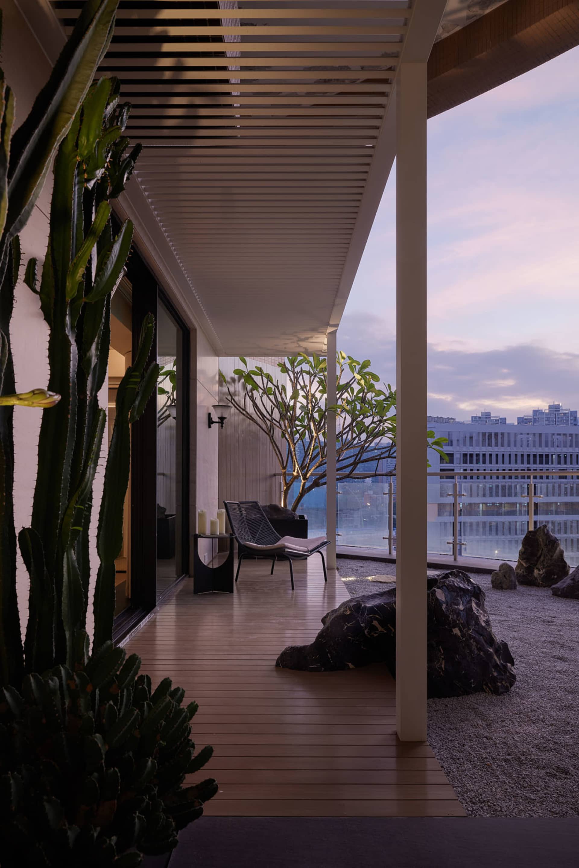 A modern residential veranda featuring wooden flooring, a lounge chair, plants, and stone landscaping against a backdrop of u