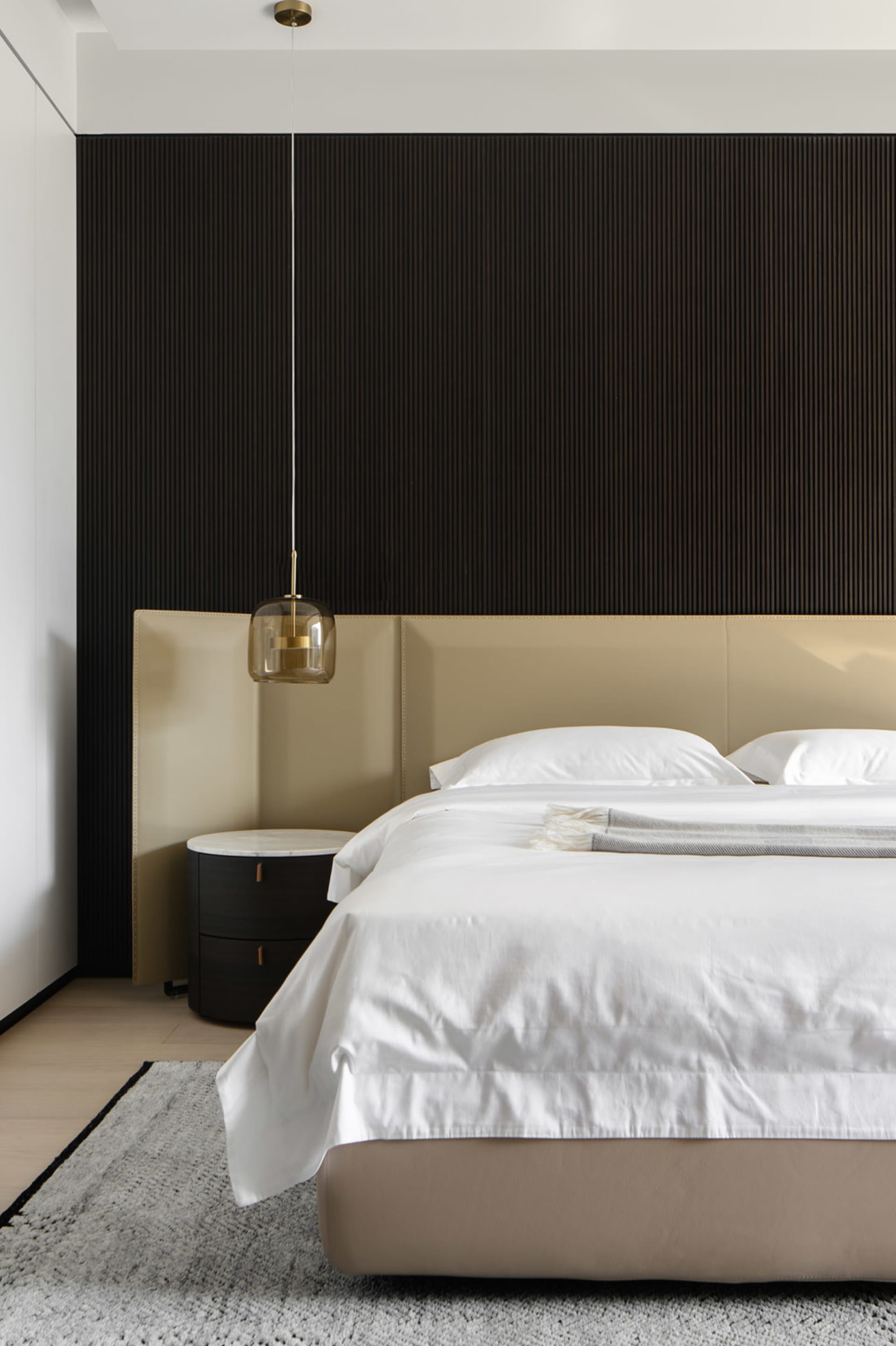 A modern minimalist bedroom with a beige headboard, white bedding, and a dark wood accent wall.