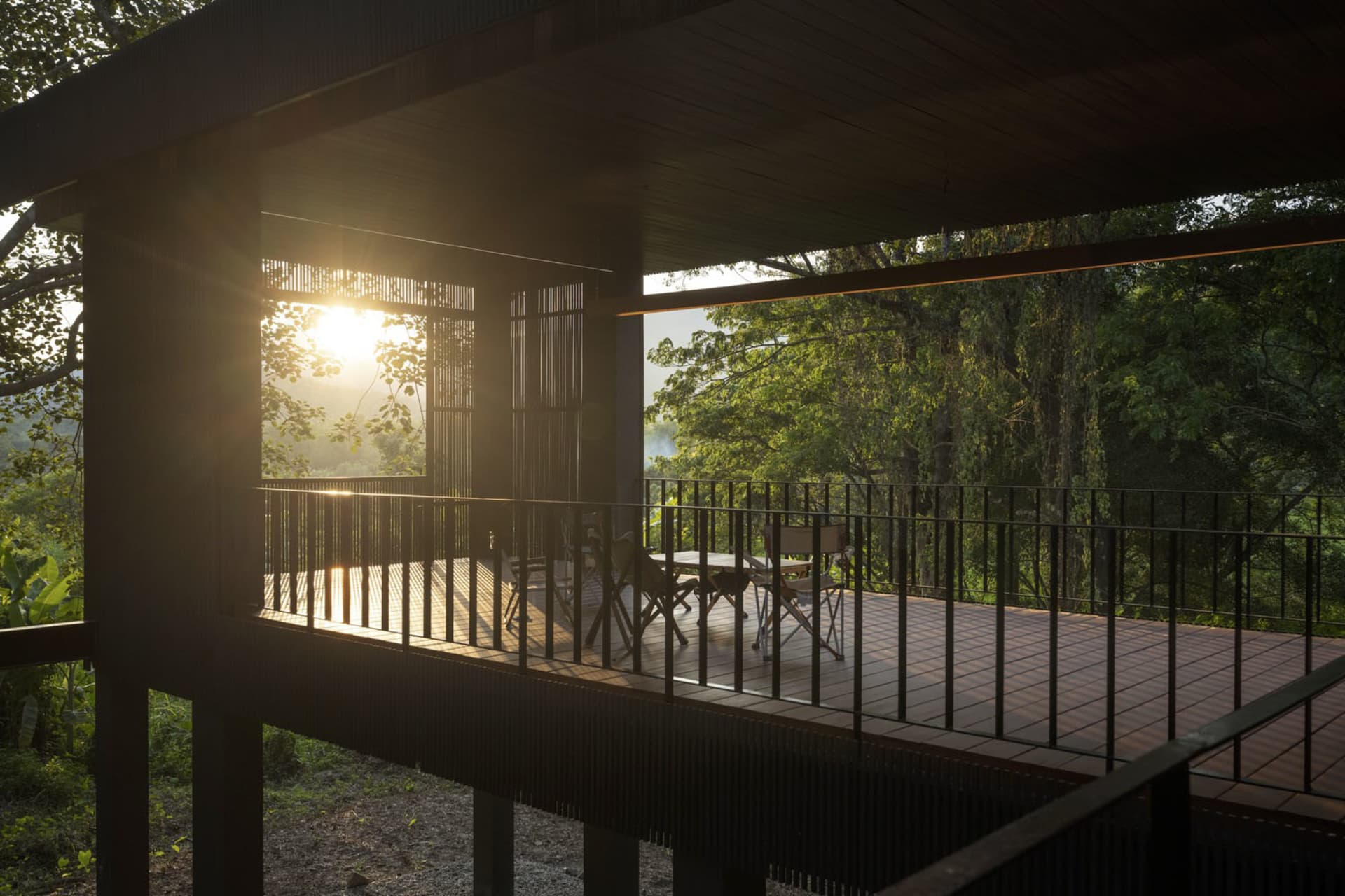 A wooden veranda with a table and chairs under a darkened roof, surrounded by lush greenery, with sunlight illuminating the s