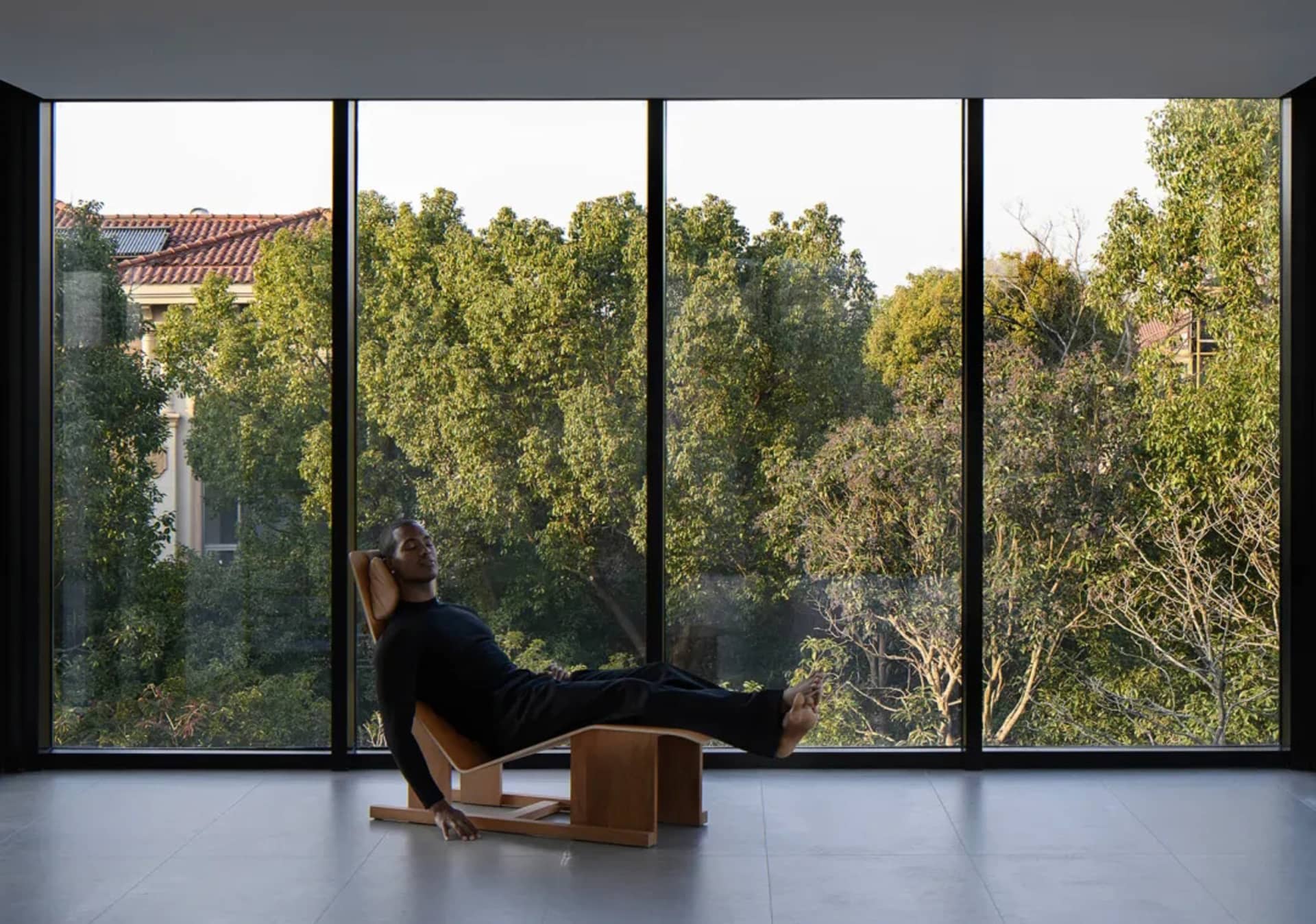 A man lying on a wooden relaxation chair in front of large windows with a view of trees.