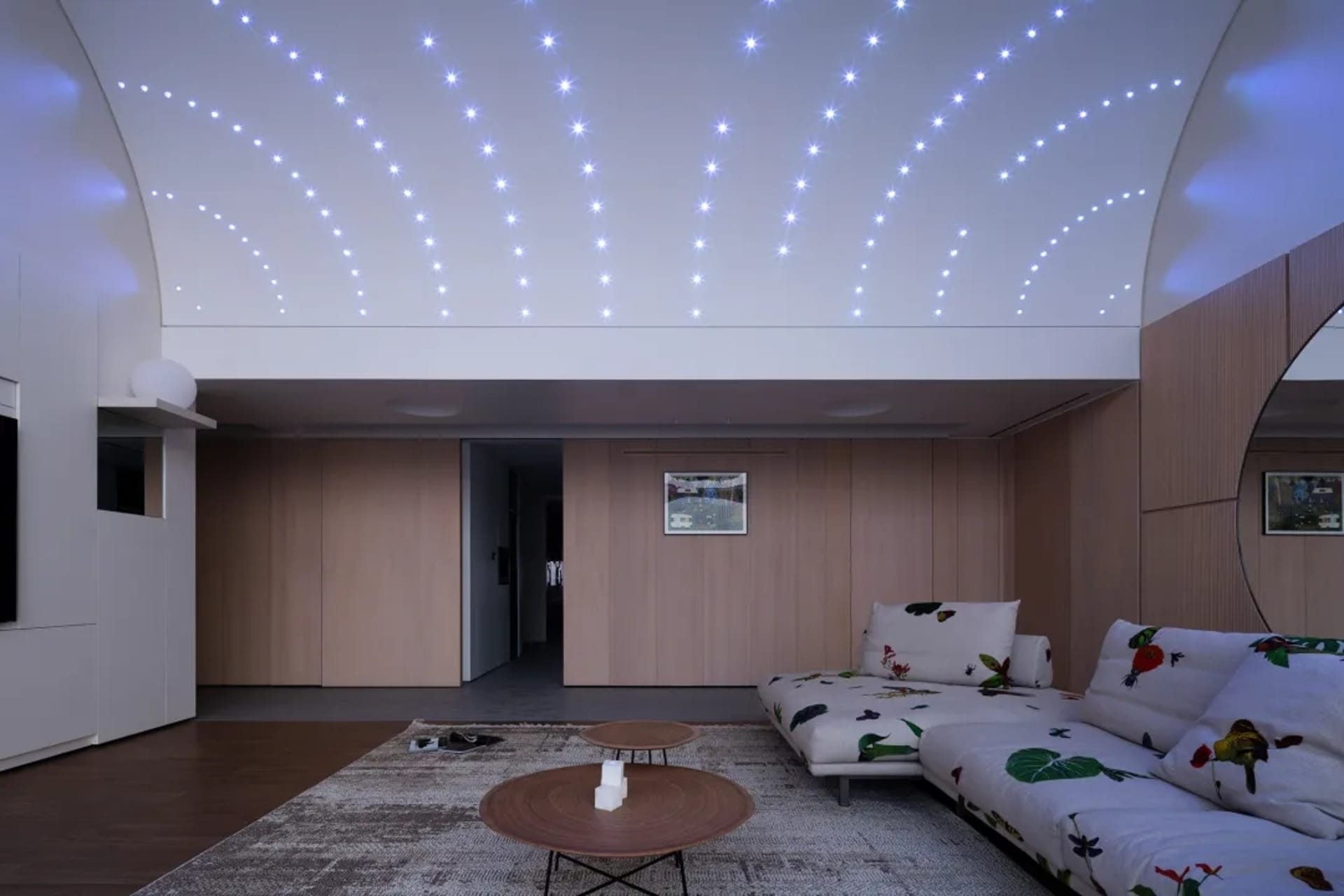 A modern minimalist living room featuring wooden walls and a white couch with floral print. The ceiling is illuminated by sta