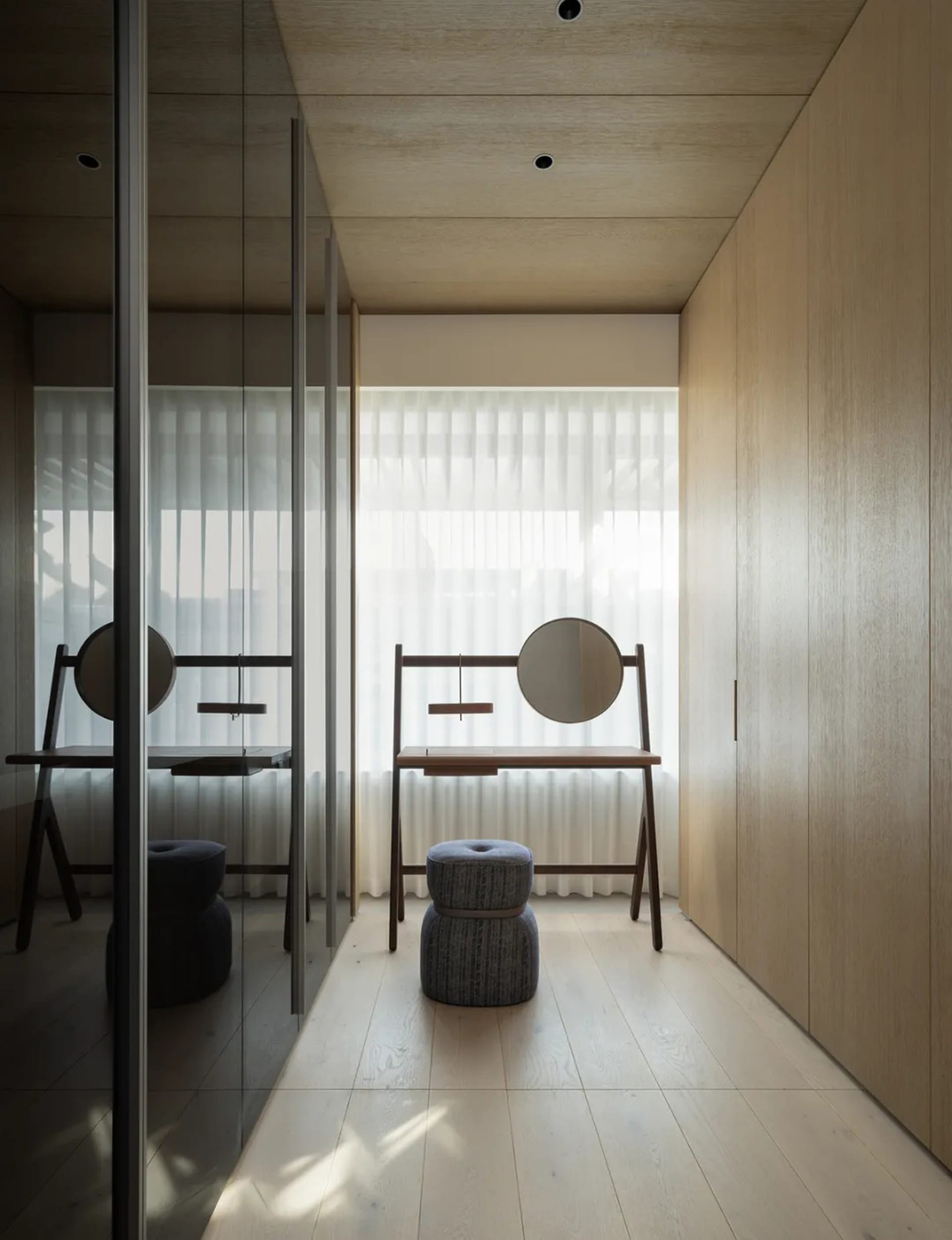 Minimalist dressing room featuring a wooden vanity with a circular mirror, a cushioned stool, and sheer curtains against ligh