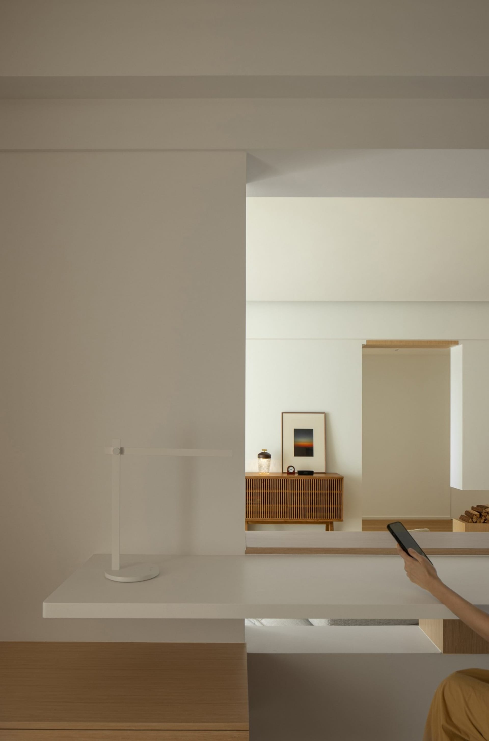 White minimalist room with a desk lamp and a person holding a smartphone.