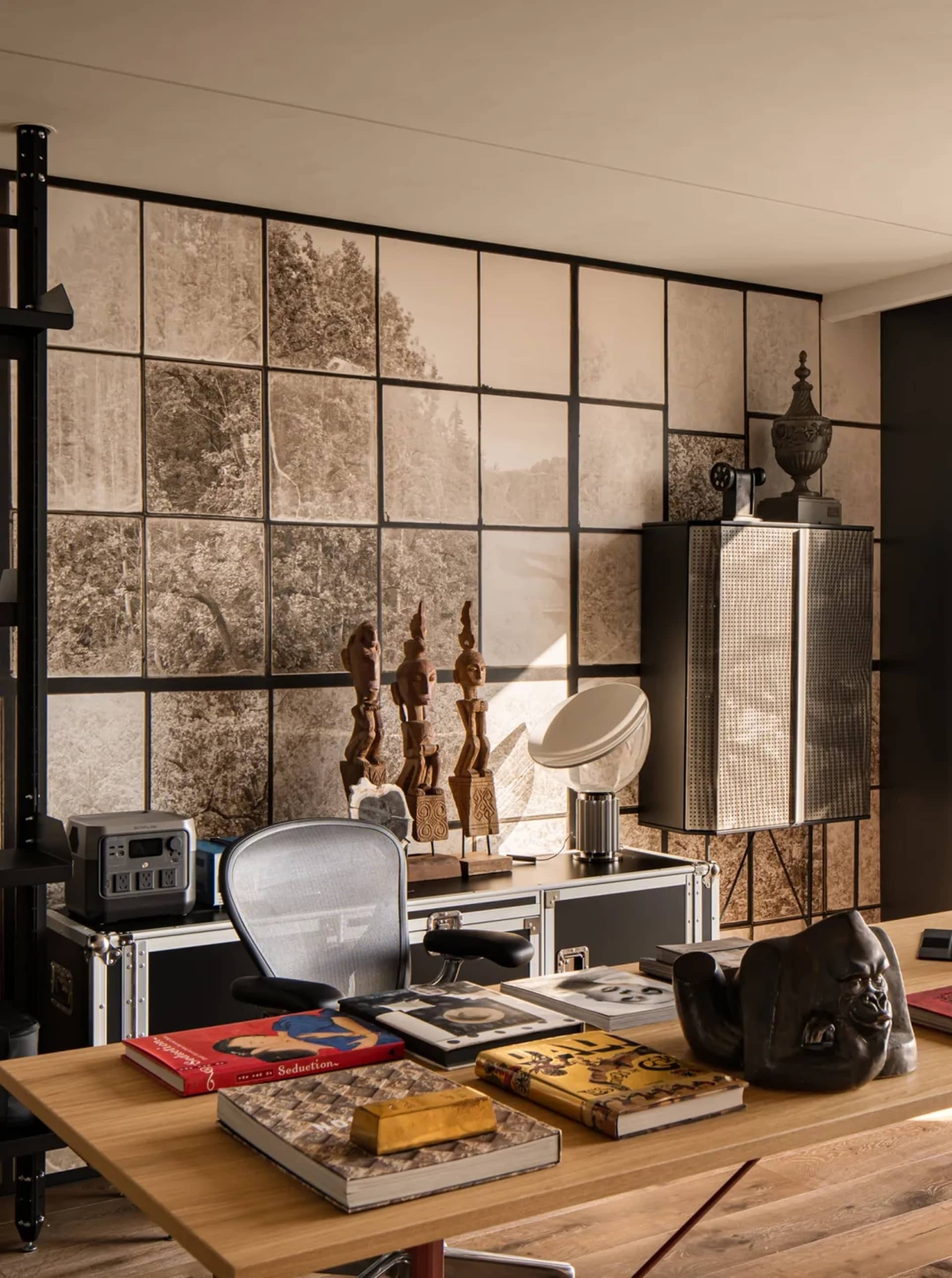 A modern, industrial-style office space featuring a wooden desk, metal shelving, and glass windows with a view of trees.
