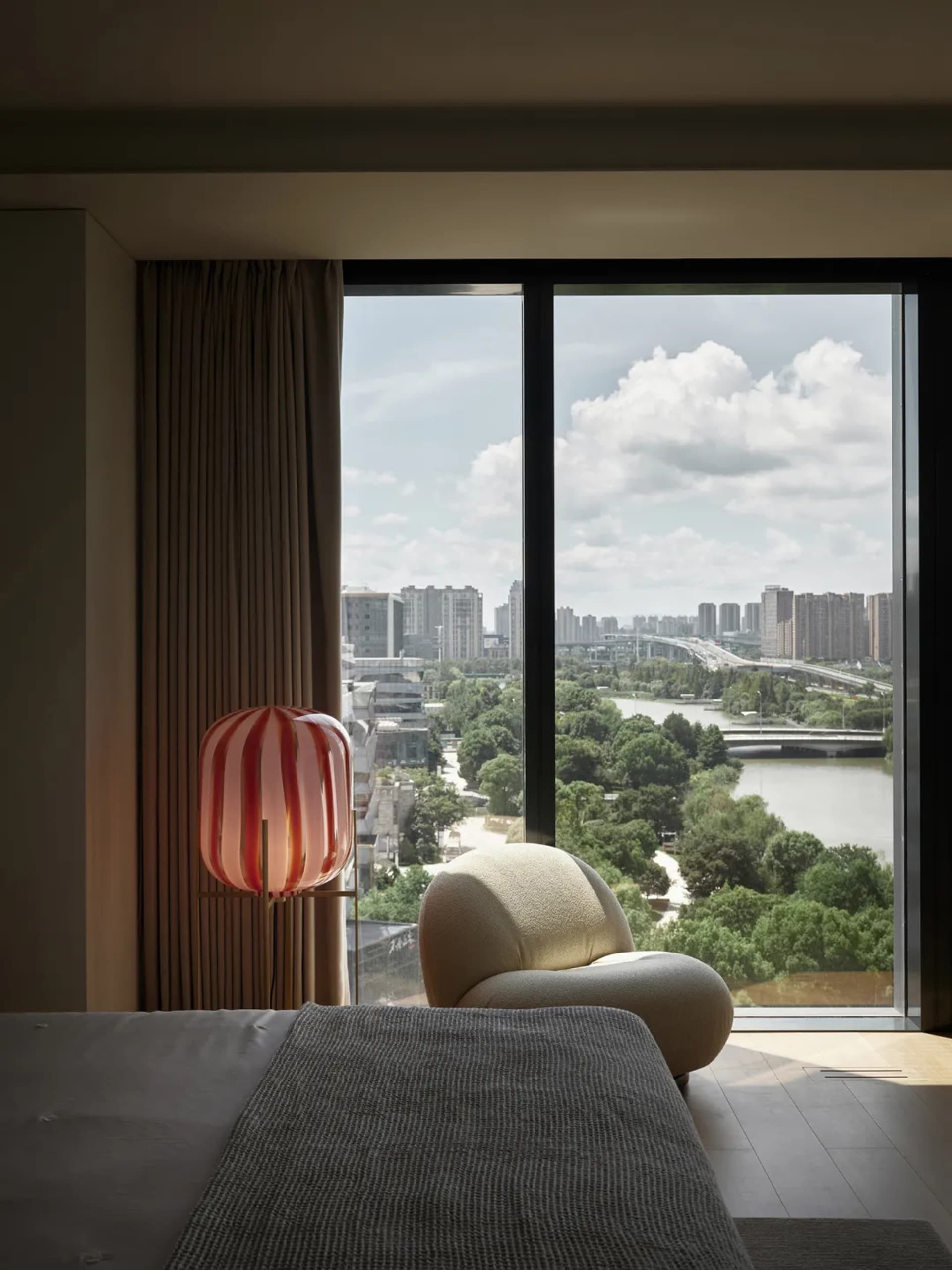A modern view lounge with a textile chair, lamp, and window overlooking a cityscape and river.