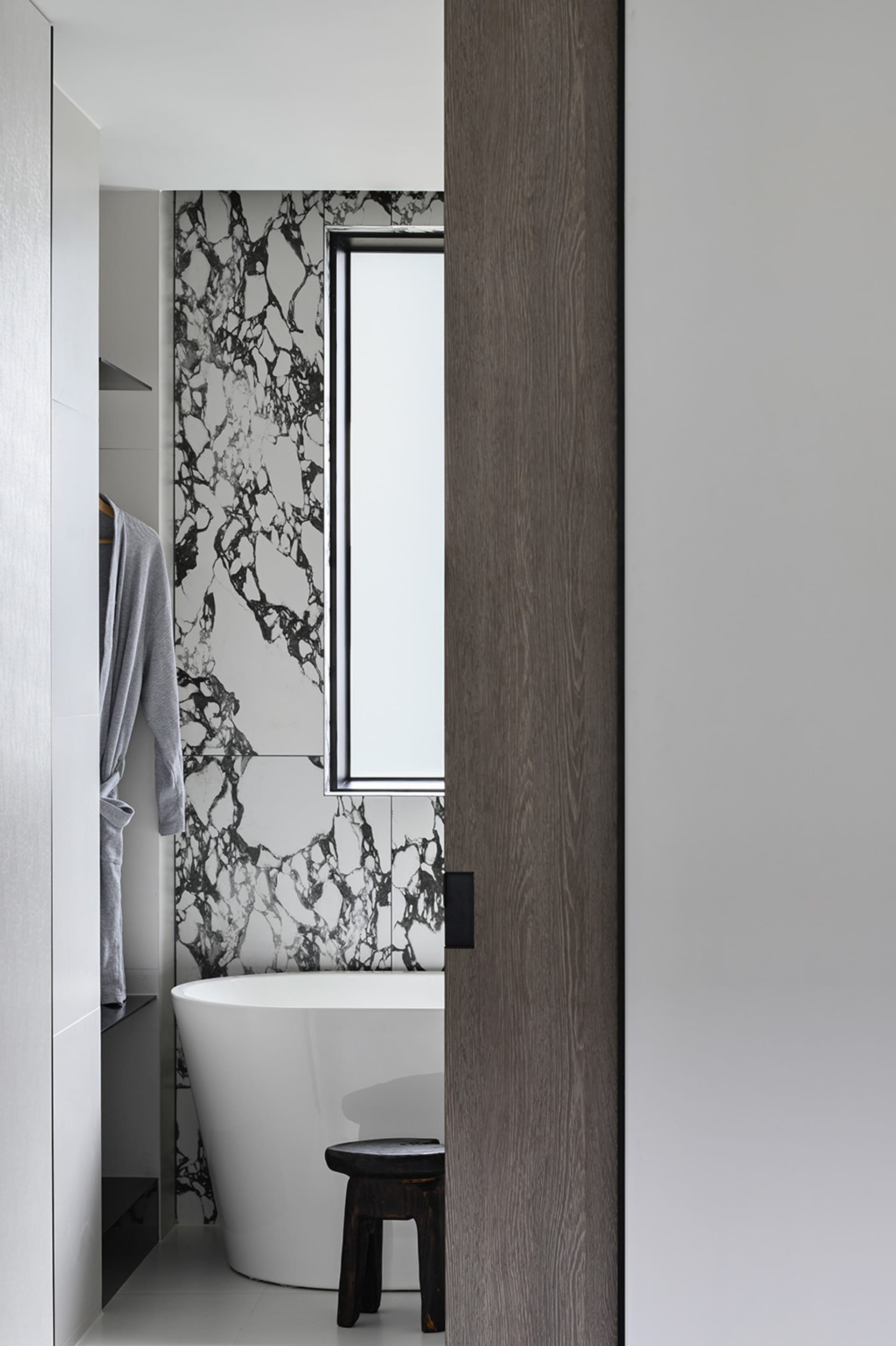 A minimalist bathroom featuring a white bathtub with a marble accent wall, a wooden stool, and a grey robe hanging