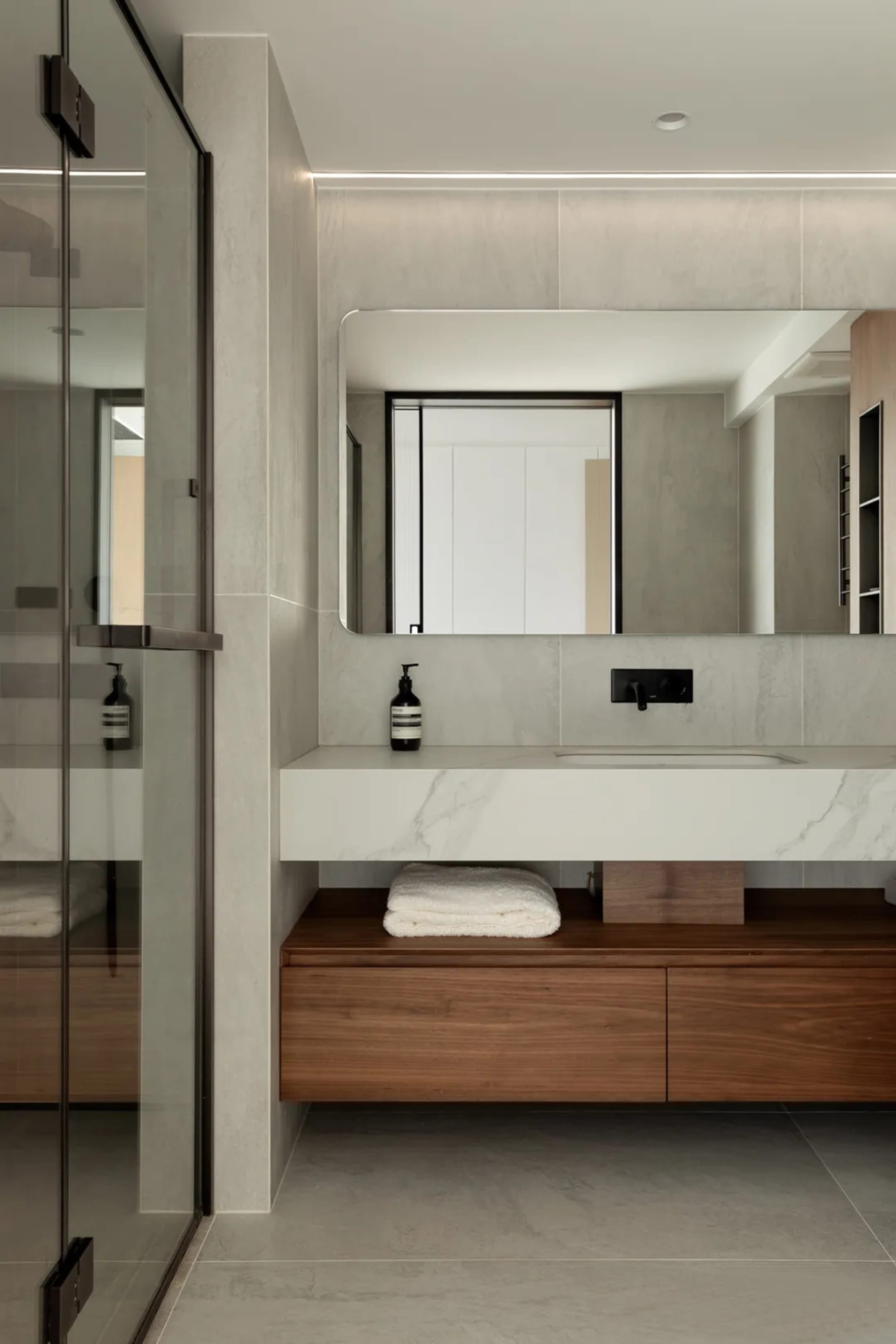 Modern minimalist bathroom with marble countertop and wooden storage, featuring a sleek sink, large mirror, and glass shower