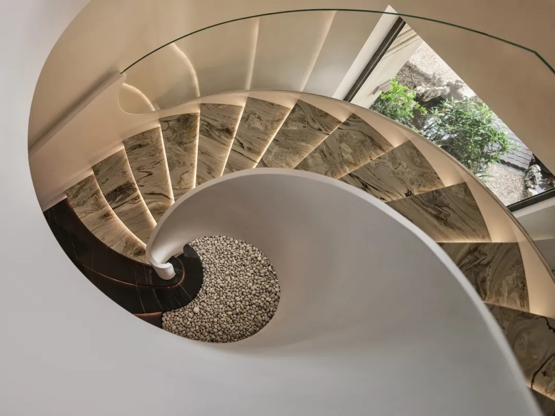 A modern spiral staircase featuring marble steps and glass railings with a white wall and green foliage in the background.