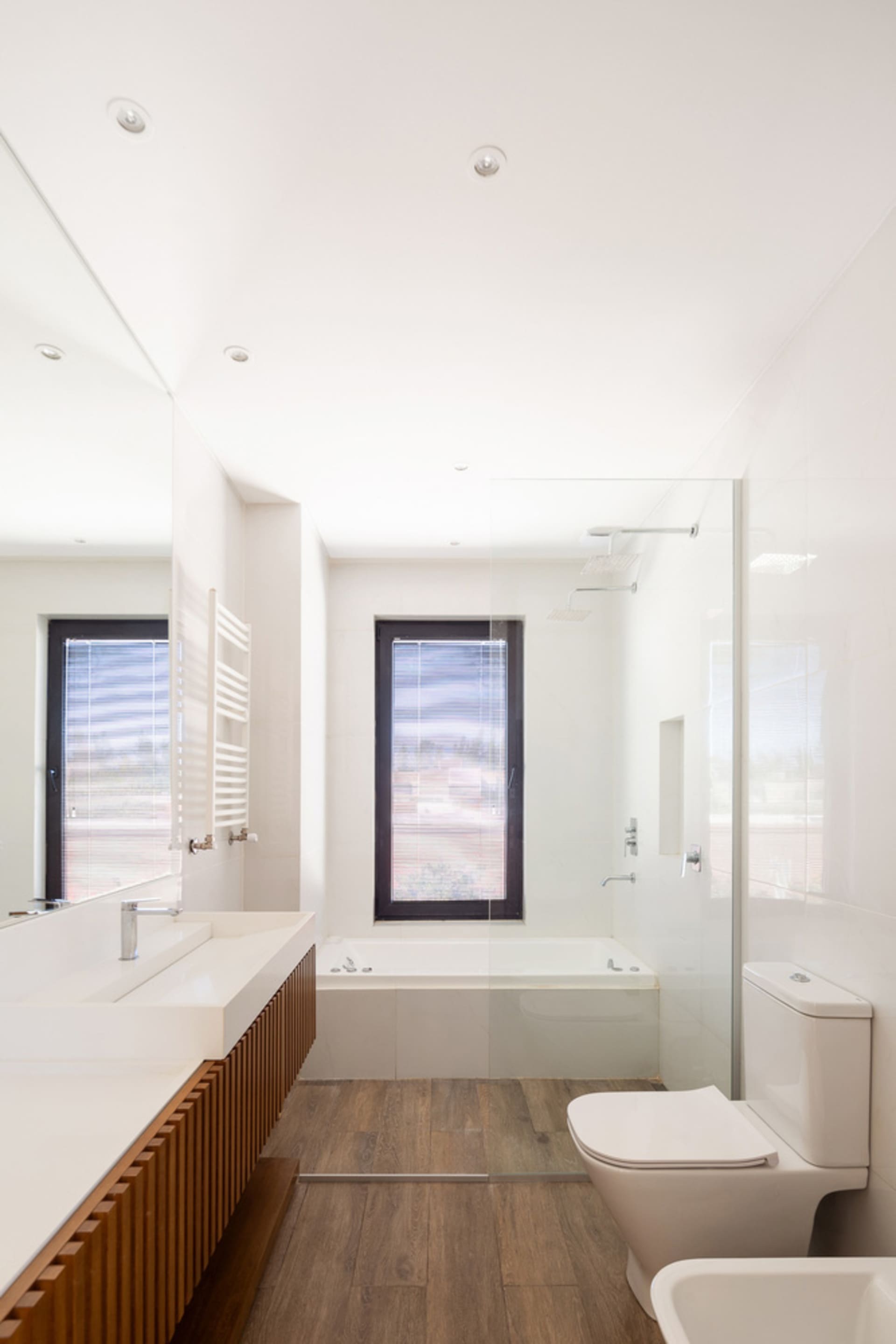 Modern bathroom with white tiled walls, a wooden floor, a glass shower enclosure, a bathtub, a sink, a mirror, and a toilet.