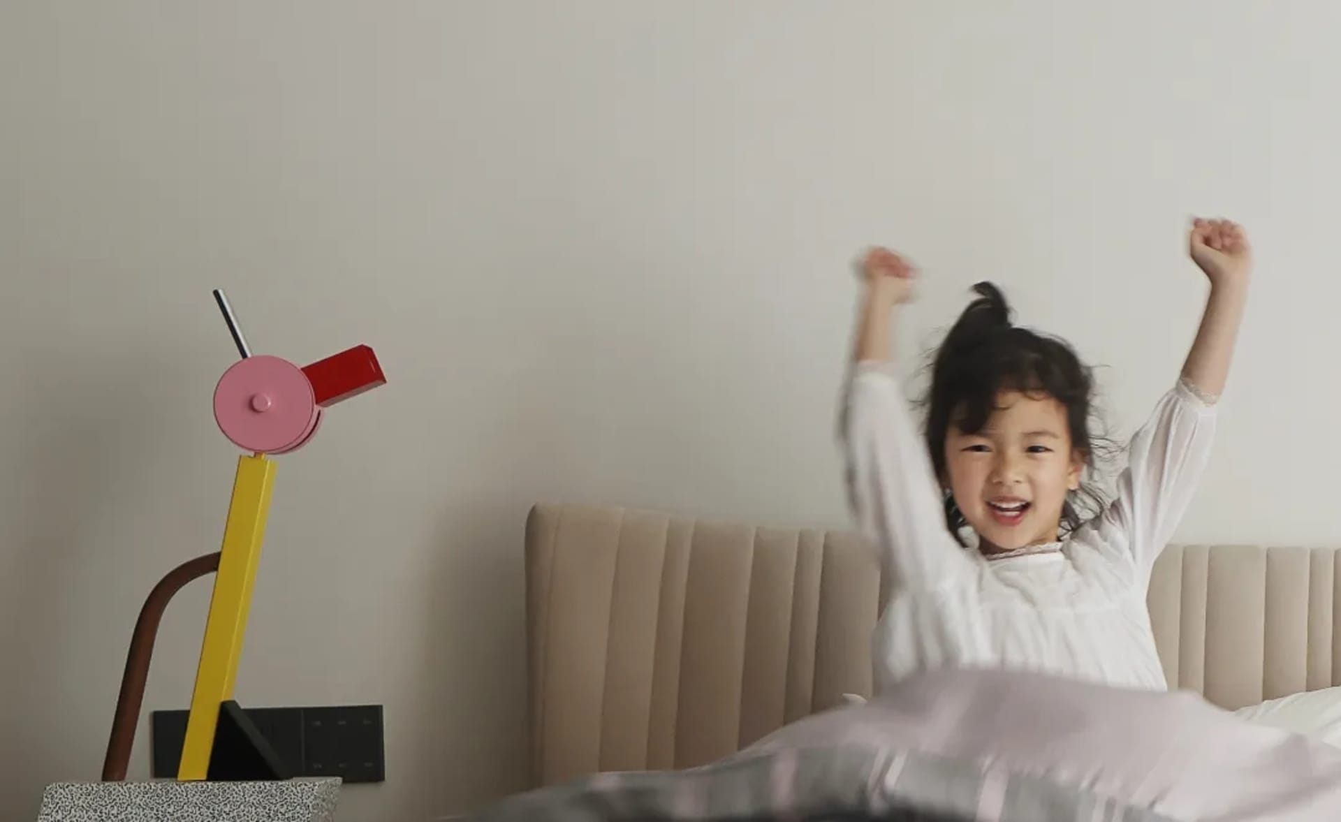 A young girl sits on a bed with her arms raised, wearing a white shirt, in a modern chic children's room with neutral furnitu