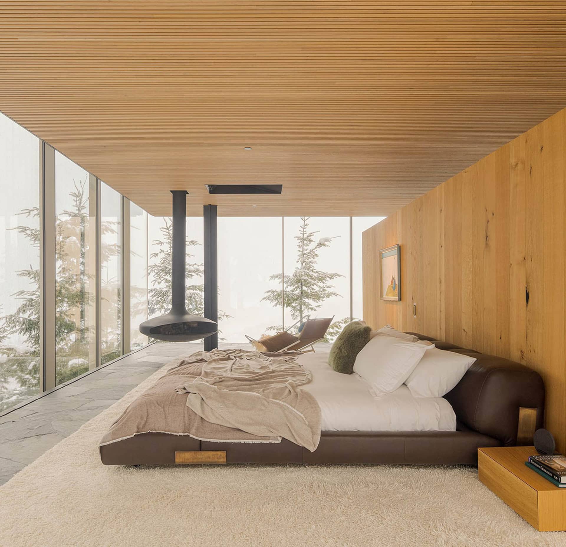 A modern bedroom featuring a leather bed with white bedding, a hanging fireplace, and a view of snowy trees through large gla