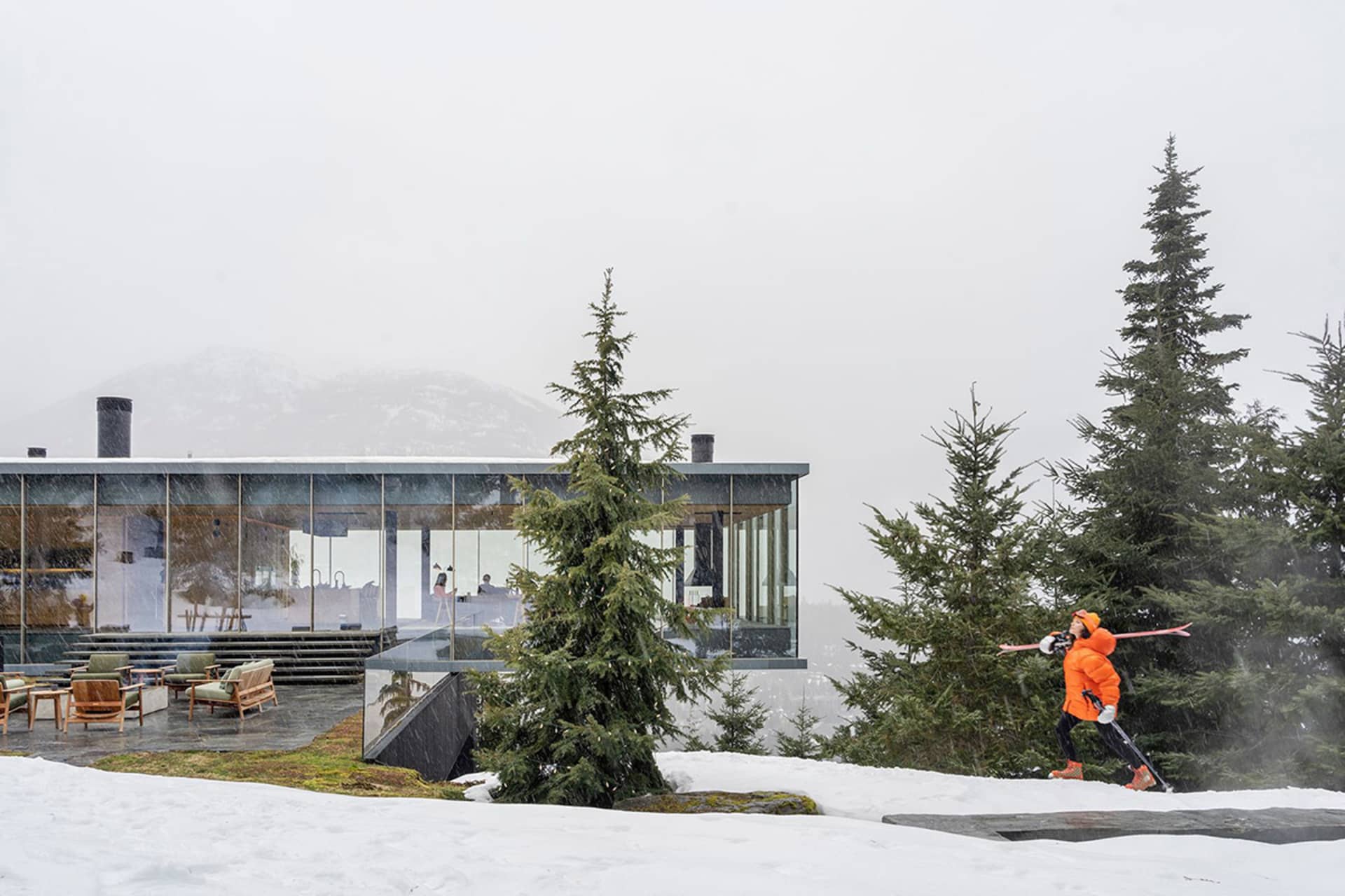 Modern residential building with glass walls and steel accents surrounded by snow and evergreen trees, with a person skiing i
